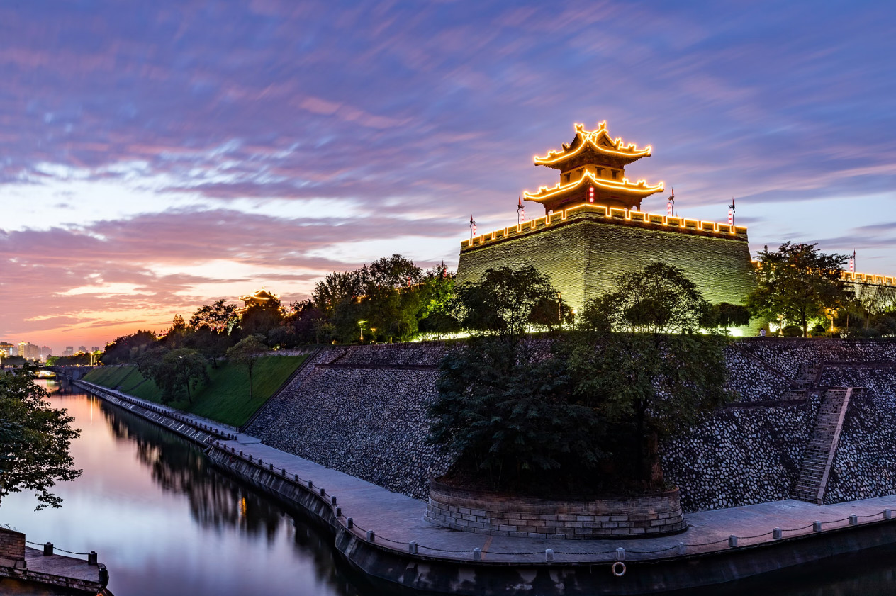 西安城牆夜景：暮色裡角樓燈光勾勒飛簷輪廓，紫粉色晚霞襯著平靜河水，是西安夜遊必打卡的古風絕美夜景