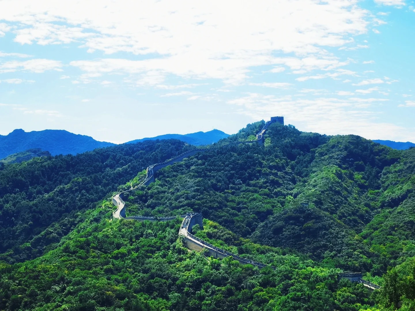 盛夏時節，八達嶺長城如一條青色巨龍，在層巒疊嶂的蒼翠林海間蜿蜒穿梭，直至盤踞峰頂的烽火臺，澄澈藍天與細碎流雲為這幅壯闊景緻更添高遠之美。