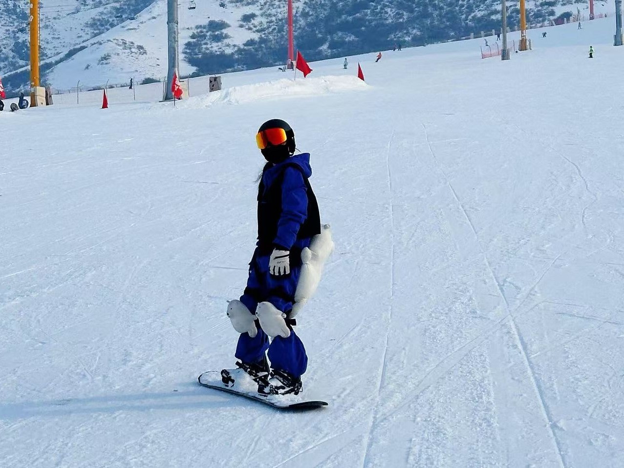 平整機壓雪道上，藍黑連體滑雪服的雪友腳踩單板平穩滑行，佩戴黑頭盔與炫彩護目鏡，搭配可愛卡通護臀護膝。場地插著紅旗與防護樁，雪道寬闊空曠，穿搭童趣滿滿，盡顯休閒滑雪狀態