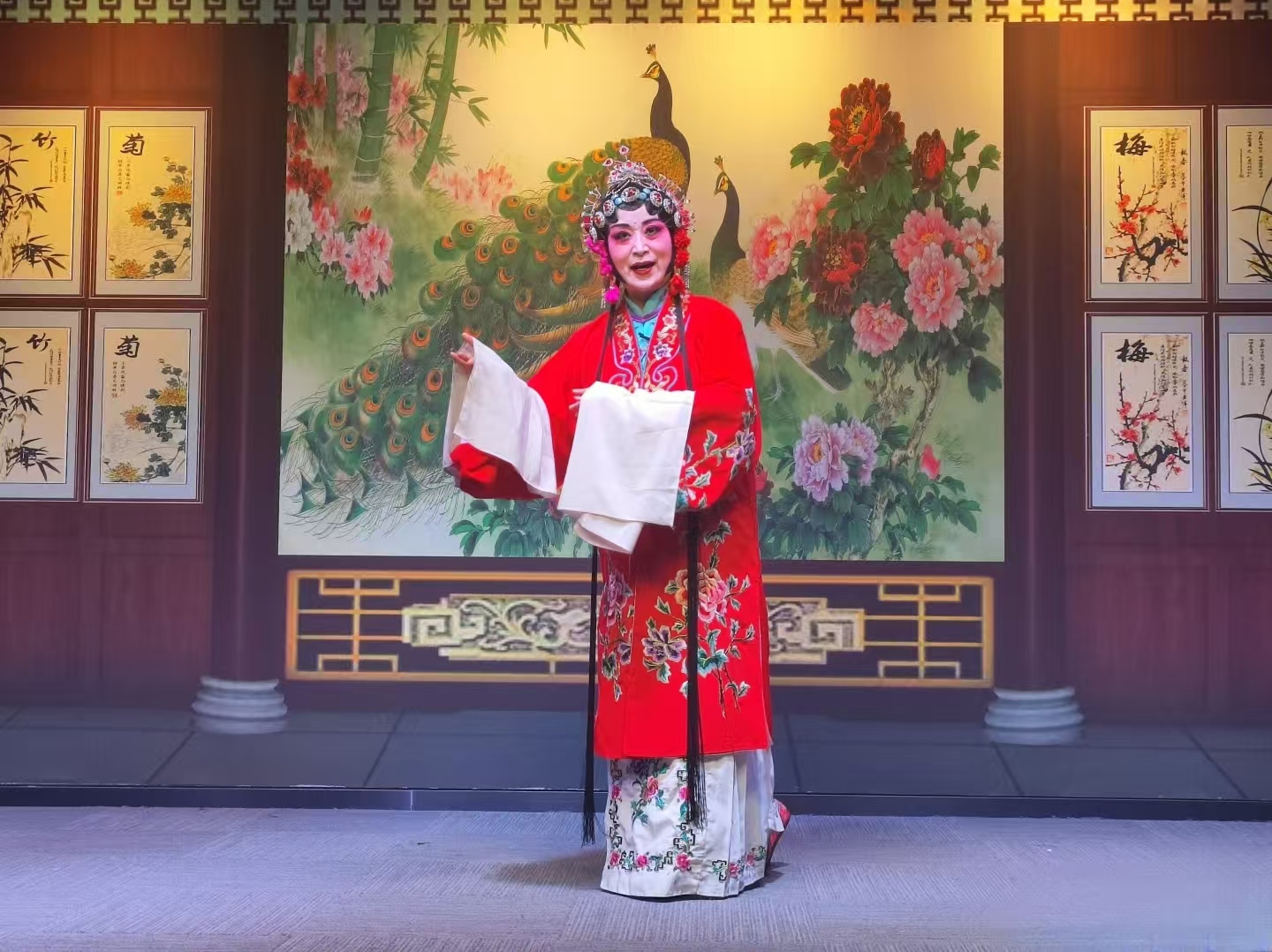 戲曲演員著大紅繡花鳥戲服，頭飾華麗，於繪有孔雀牡丹的背景前展袖表演，盡顯戲曲的明豔與典雅