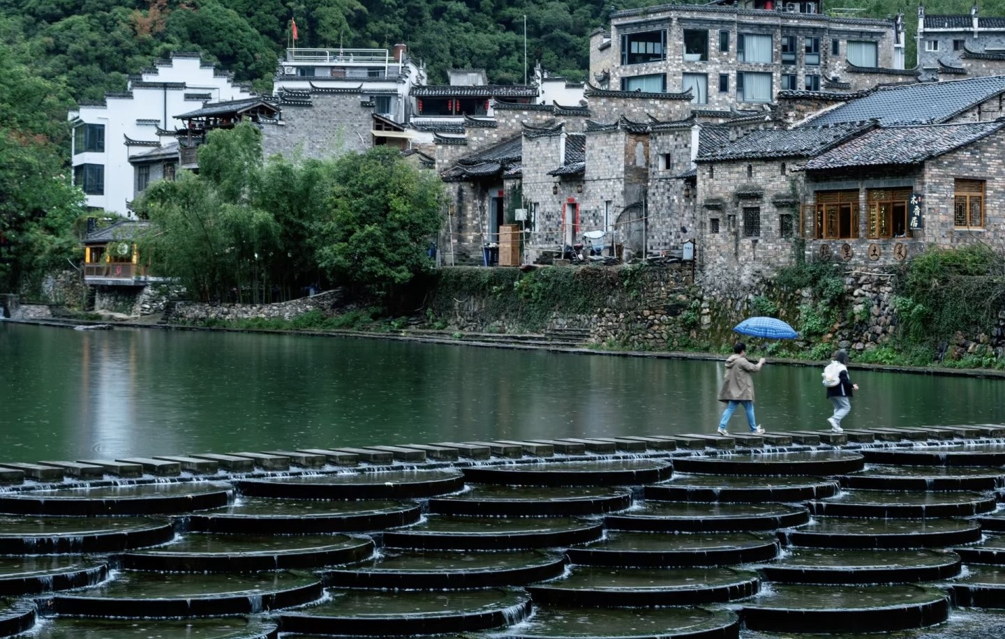 細雨中的古村被一層薄霧籠罩，墨色的石板橋橫跨在碧綠的河面上，撐著藍傘的行人緩步走過