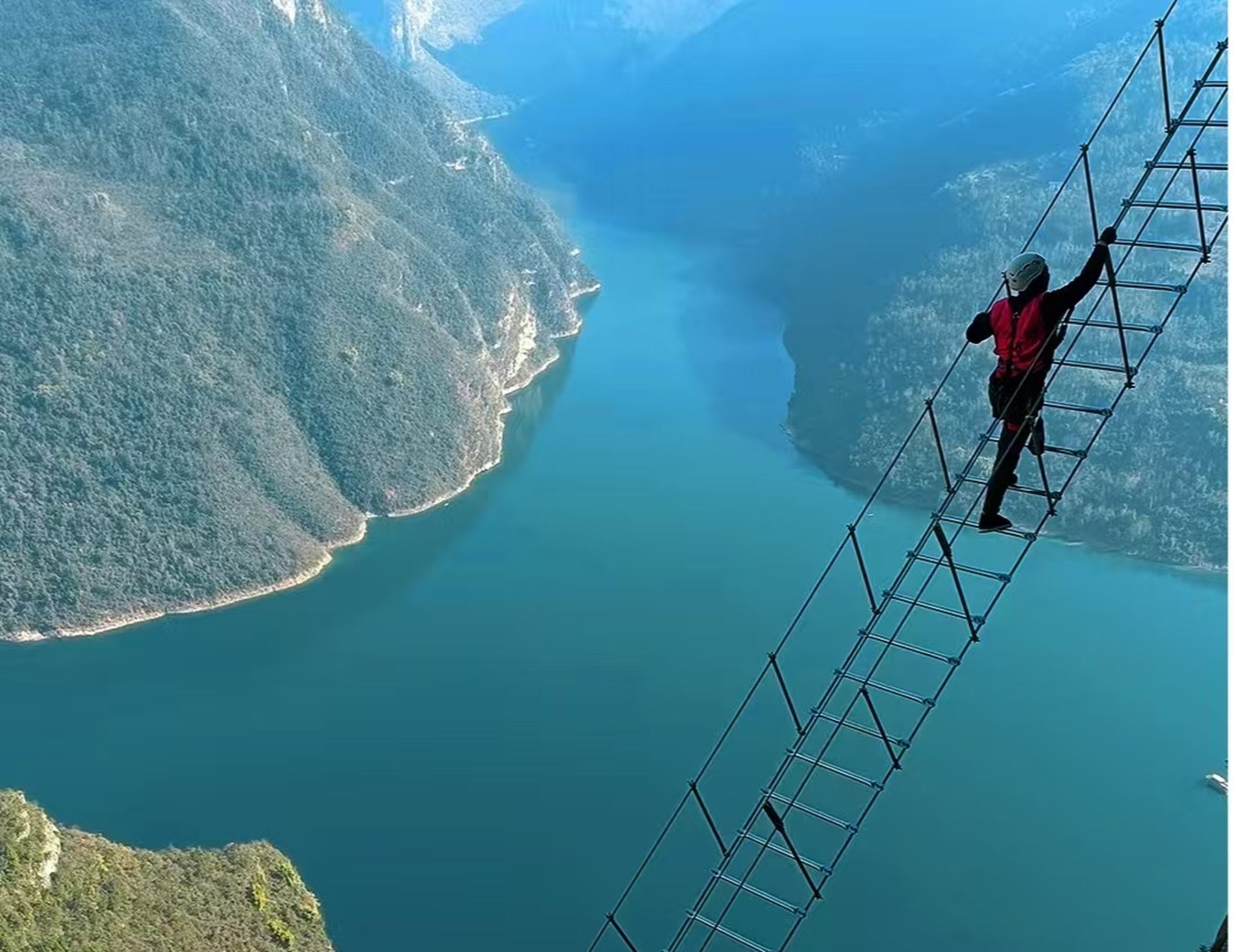 紅衣者在峽谷高空金屬梯上前行，青綠江面繞著翠嶺，懸空步道與山河相映，步伐裡裹著輕極限的刺激與壯闊觀景體驗