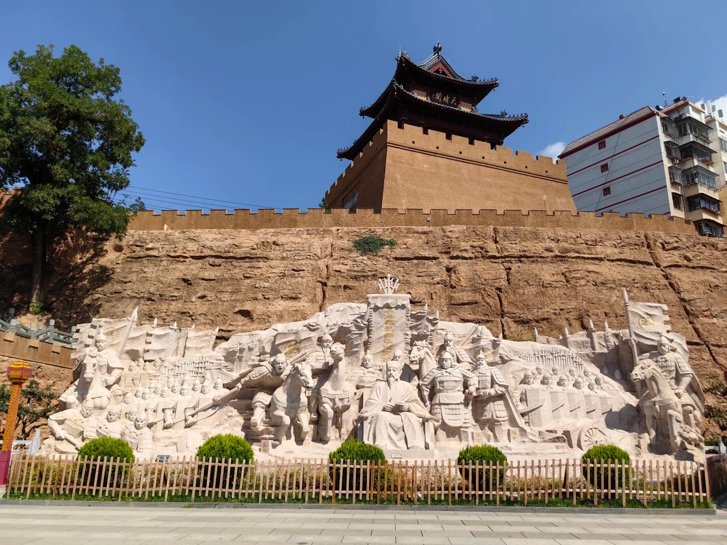 這是甘肅天水伏羲城景觀中的大型歷史浮雕牆。畫面中央，巨幅石質浮雕以磅礴氣勢鑲嵌於古城牆基座，雕刻群像動態豐富，生動再現了上古傳說或重大歷史場景，人物衣袂飛揚、戰馬奔騰的細節彰顯高超的雕刻技藝。浮雕上方，夯土城牆與重簷歇山頂城樓古樸厚重，黃土色牆體鐫刻著歲月痕跡，展現明代軍事防禦建築特色。城牆一側，現代高層建築悄然入鏡，形成古今對話的獨特畫面。作為天水歷史文化名城的核心景觀，這裡將神話傳說、軍事文明與城市發展熔鑄一體，成為傳承伏羲文化、展現隴原歷史的重要載體。