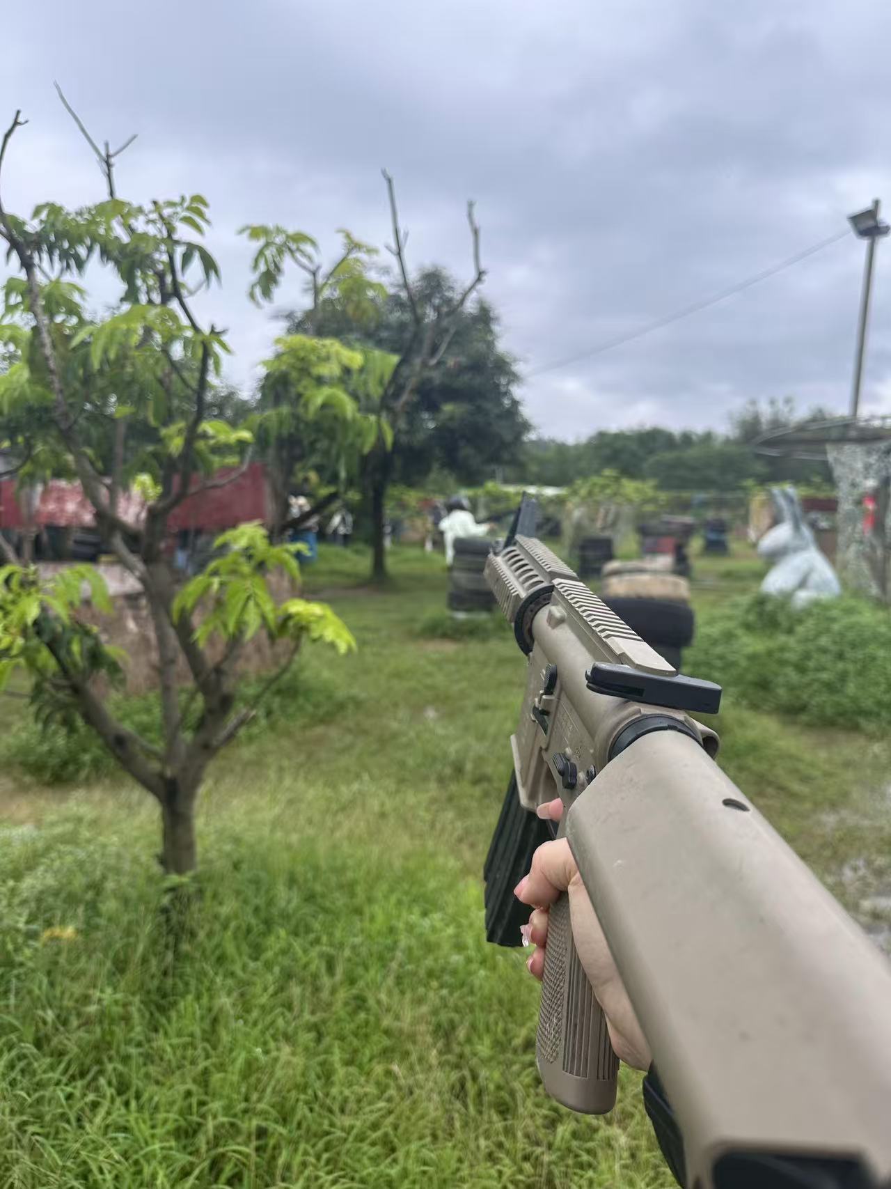 第一視角沉浸式 CS 戰場！手持沙色仿真槍，瞄準前方叢林陣地，草地、樹木與掩體環繞，彷彿下一秒就要開啟攻防戰，代入感拉滿