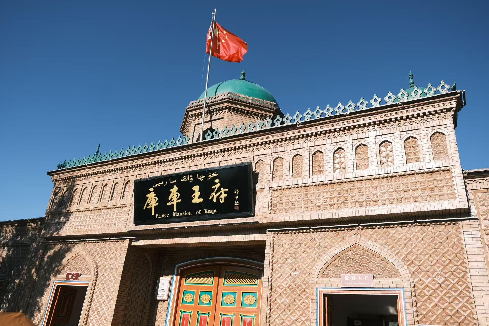 庫車王府的建築在晴空下格外醒目，土黃色磚雕牆面紋理精緻，綠色穹頂與彩繪木門盡顯西域風情。門楣上的漢、維雙語牌匾莊重典雅，五星紅旗迎風舒展，訴說著百年曆史的厚重與新生