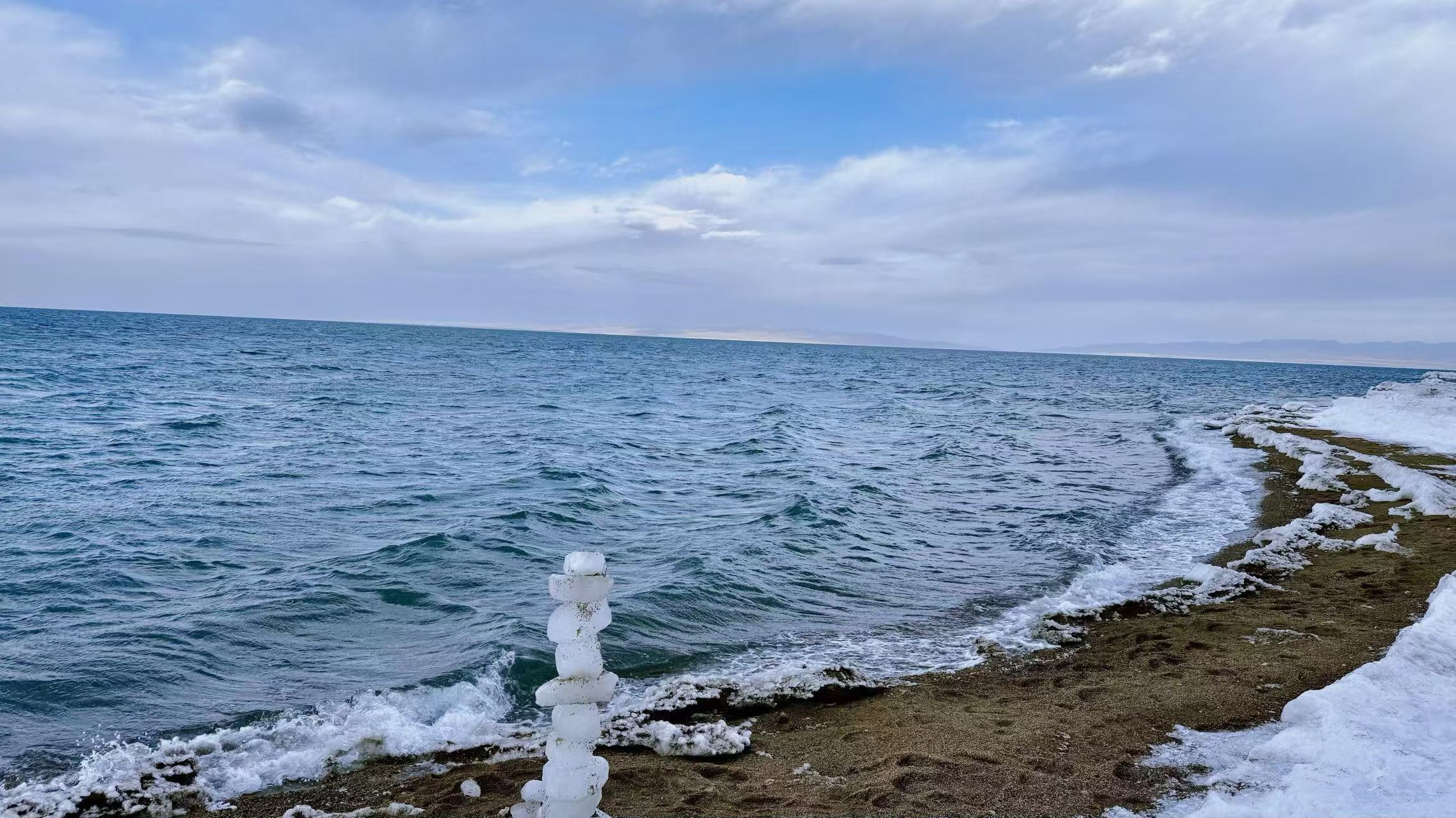 湖邊冰瑪尼靜靜矗立，湖水翻湧層層碧波，浪花輕拍岸邊殘冰。雲天遼闊，寒風漫過湖面，帶著高原獨有的清冷與安寧