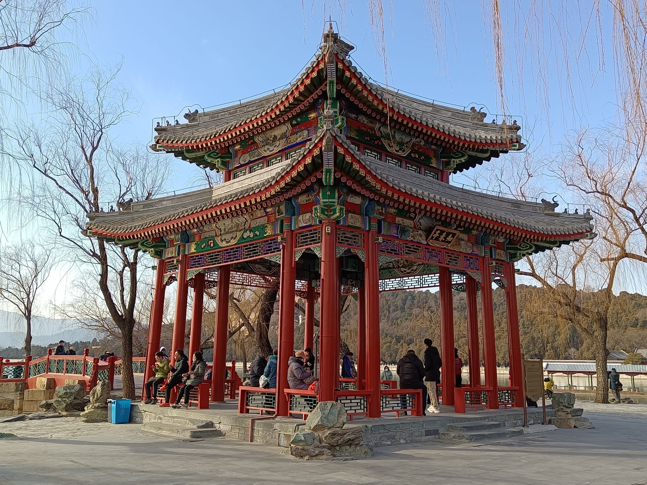 頤和園知春亭立於湖畔，重簷攢尖頂造型典雅，紅柱灰瓦在金黃柳絲間格外亮眼，亭中游人休憩，盡顯秋日園林的靜謐雅緻