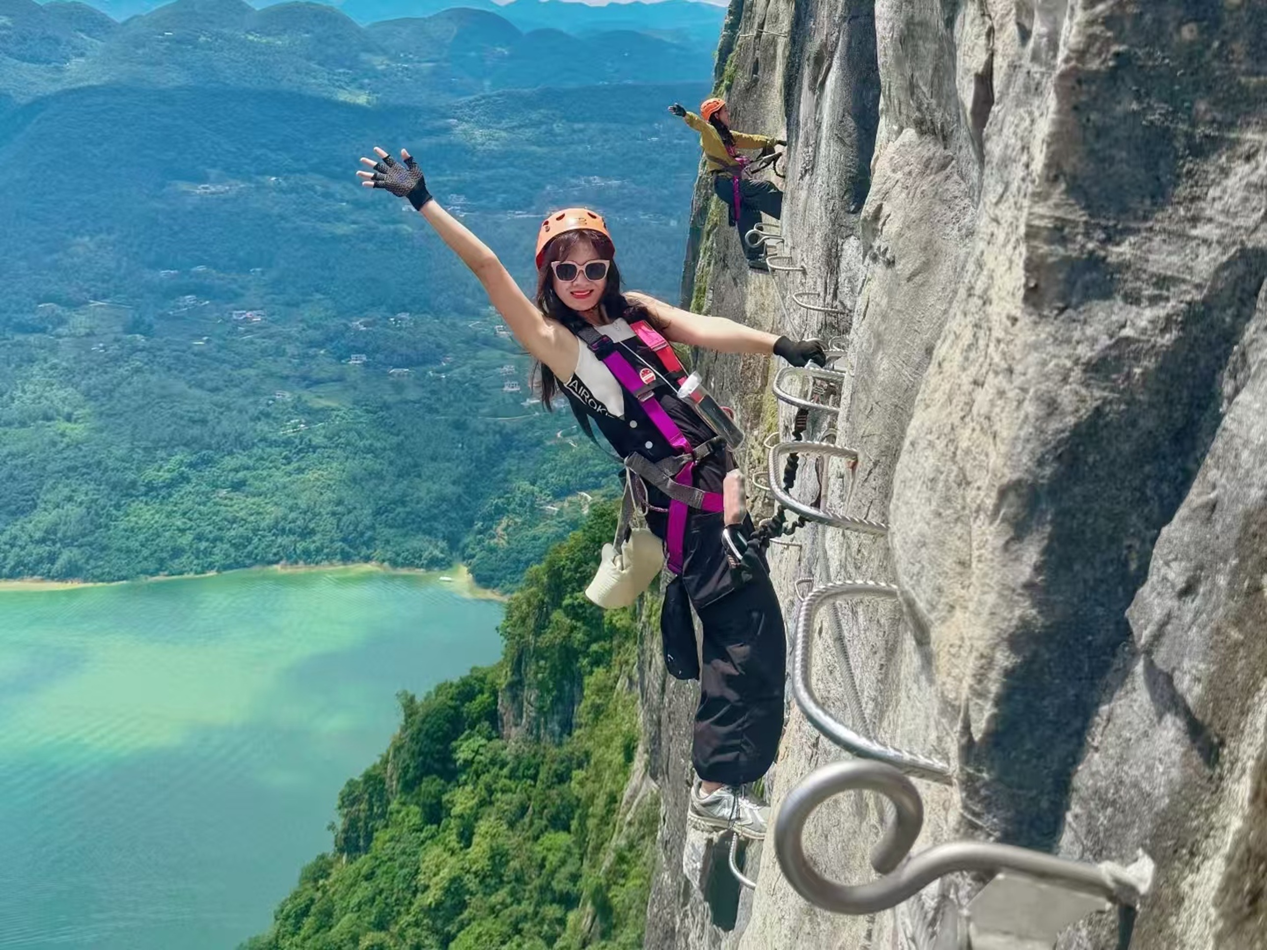 橙盔女子在崖壁梯上展臂，身後同伴同框，碧江襯著翠嶺，笑意裡裹著高空探險的鮮活與暢快