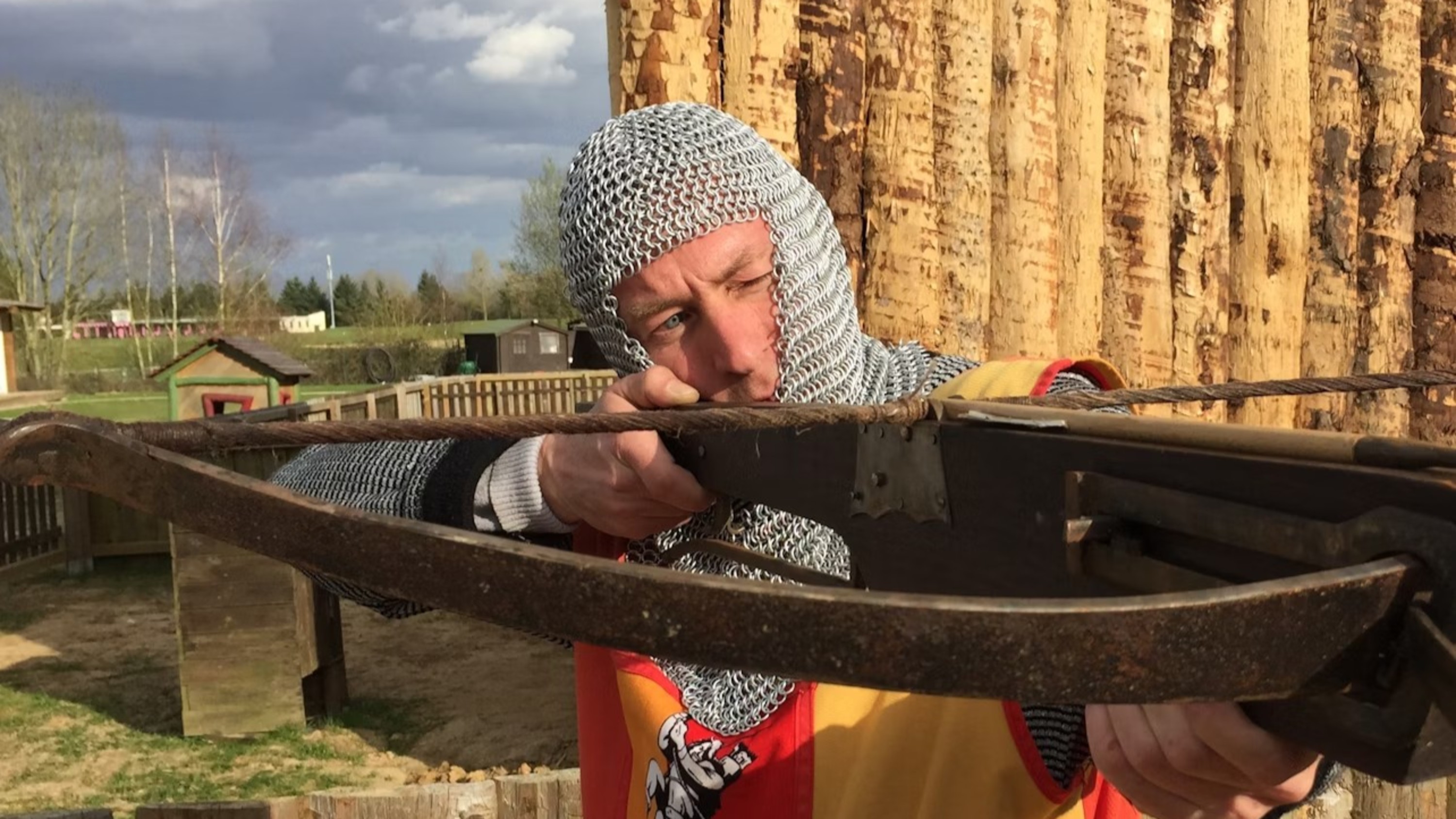 Readying his aim with a historical crossbow, this individual in chainmail appears focused on a target in an outdoor setting