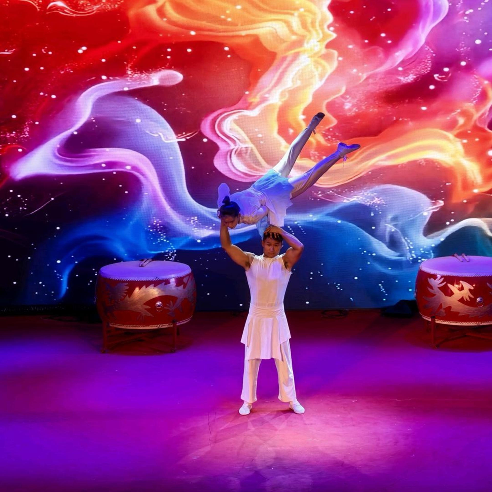 雜技表演者著白衣，男子頭頂託舉女伴，背景炫彩光影配紅鼓，動作驚險，舞臺效果震撼