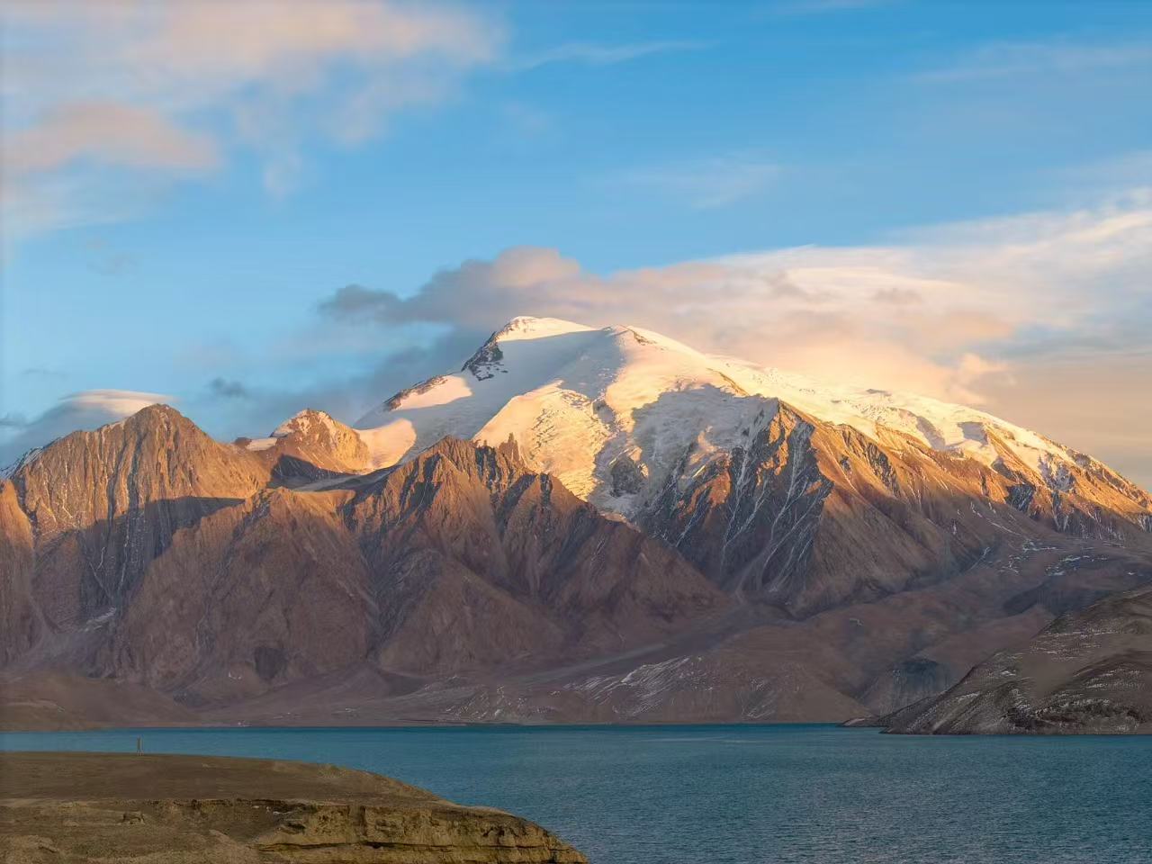 暮色中的慕士塔格峰被夕陽染成溫暖的金橙色，雪峰在餘暉中更顯巍峨。山腳下的喀拉庫勒湖泛著幽藍的波光，靜謐的水面與壯麗的山峰相映成輝