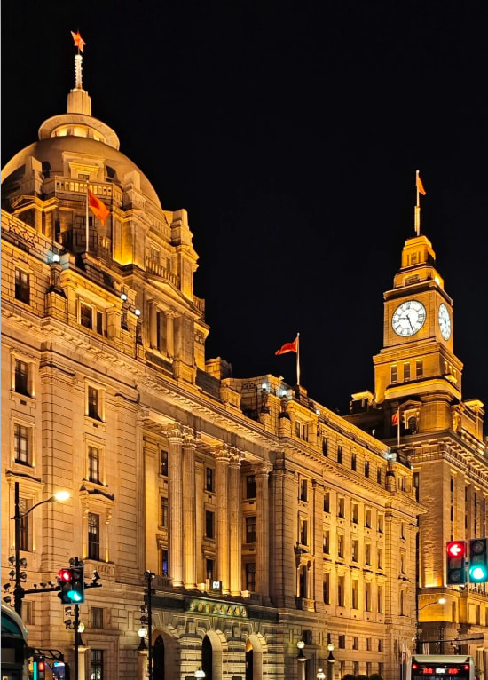 外灘核心建築夜景，米黃色石材外牆在暖光下盡顯典雅，標誌性的圓頂鐘樓與尖頂塔樓錯落有致。牆面雕刻與立柱細節清晰，紅燈籠點綴其間，整點報時的鐘樓鐘聲，訴說著外灘百年的歷史底蘊
