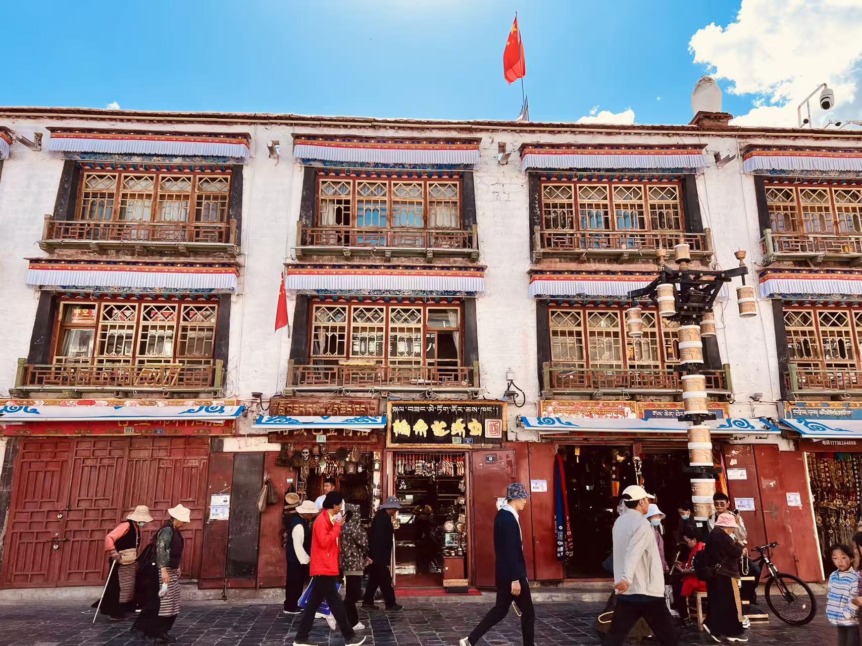 八廓街經典藏式沿街建築,白牆搭配雕花木窗與復古木陽臺,簷邊飾有彩紋布簾,樓頂飄揚五星紅旗。紅木門商鋪沿街排開,藏漢雙語招牌醒目,石板路上行人往來,滿是濃郁鮮活的拉薩市井人文氣息 八廓街經典藏式沿街建築,白牆搭配雕花木窗與復古木陽臺,簷邊飾有彩紋布簾,樓頂飄揚五星紅旗。紅木門商鋪沿街排開,藏漢雙語招牌醒目,石板路上行人往來,滿是濃郁鮮活的拉薩市井人文氣息