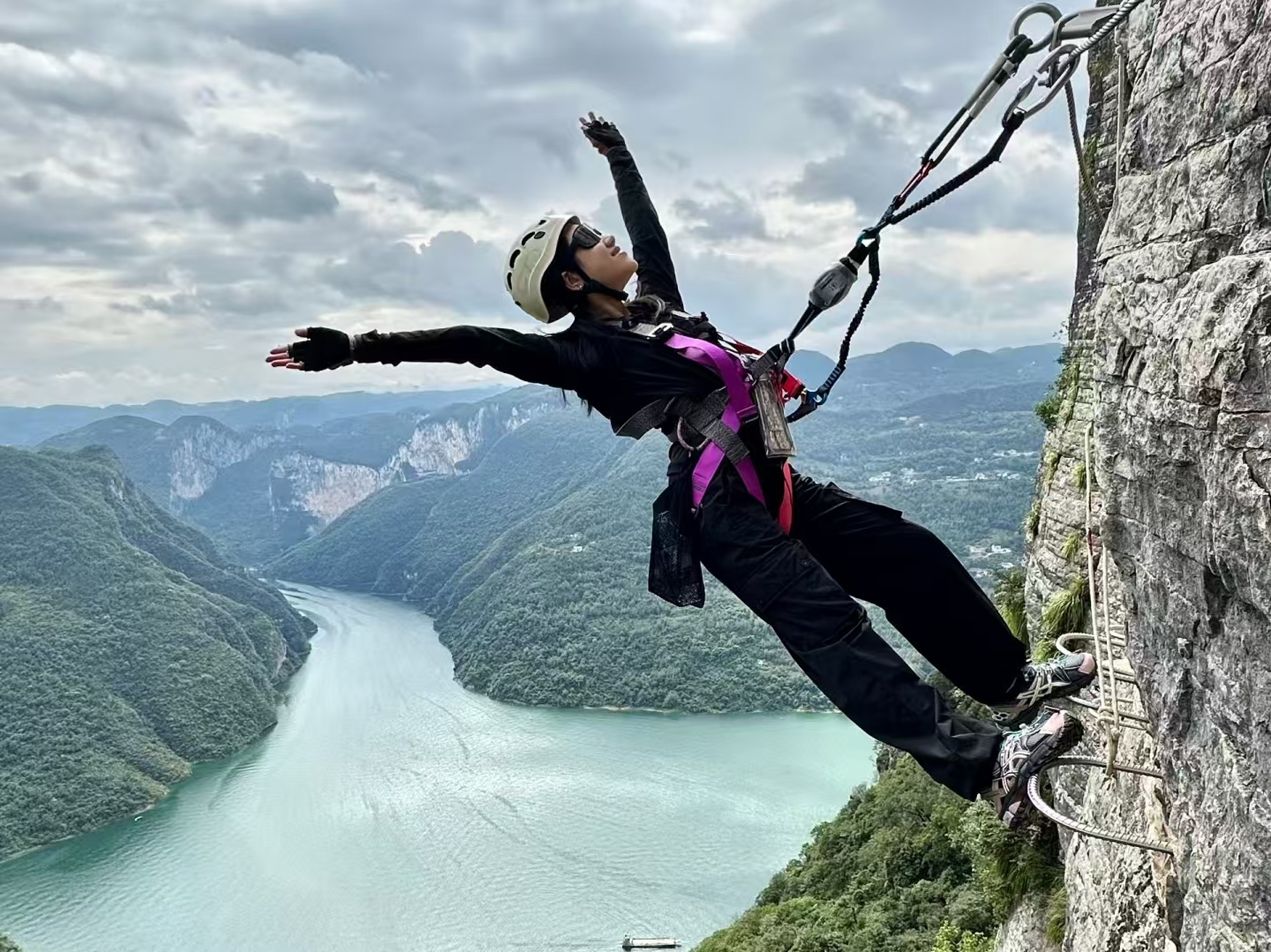 戴盔的人懸於崖壁旁，展臂後仰，江灣與層山作景，風裹著髮絲，是懸崖挑戰裡的自由與野性