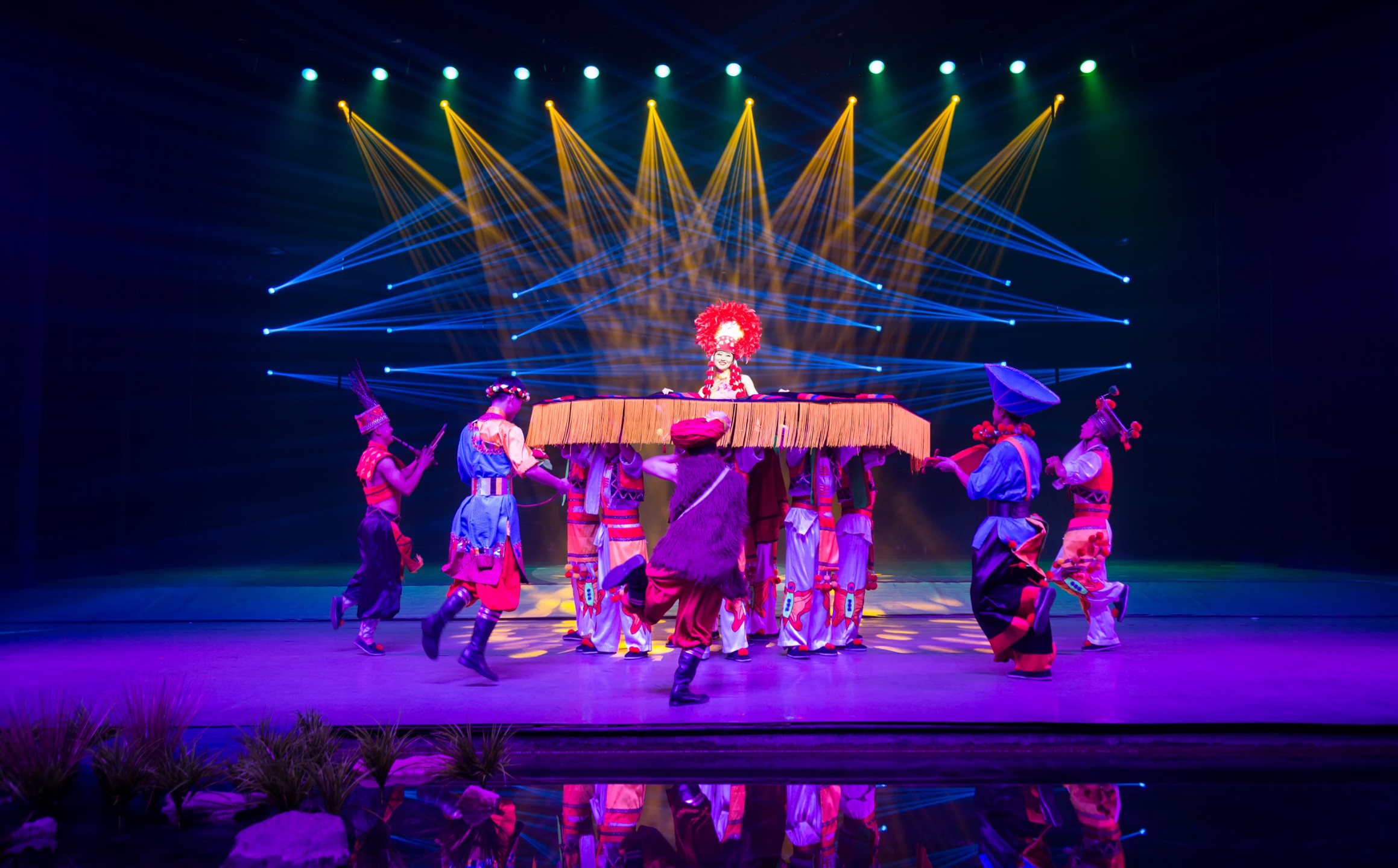 身著盛裝的主角被眾人抬在草編華蓋下，倒影在舞臺水面，演繹出濃郁的民俗儀式感