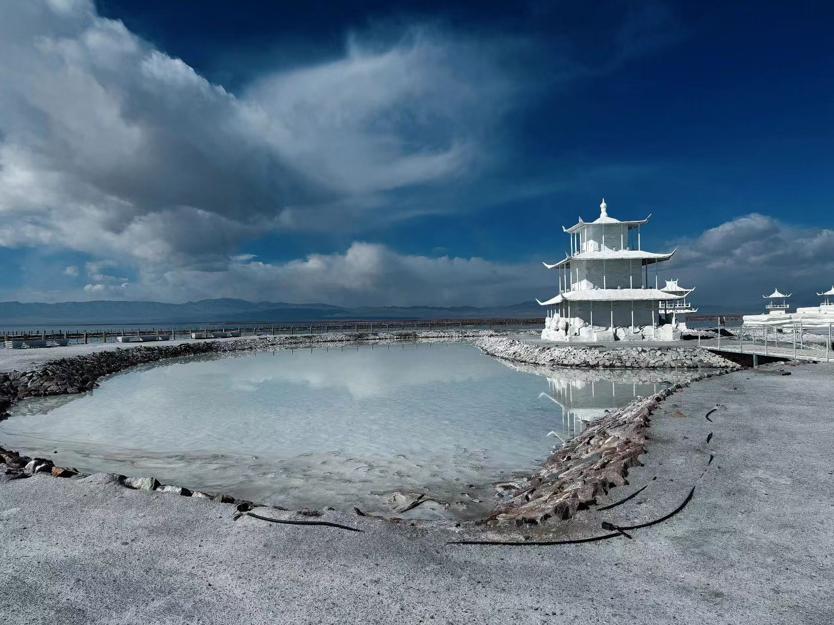 鹽湖之中坐落潔白鹽雕古亭，淺湖映著雲影，大塊流雲鋪在深藍天幕，鹽質建築別緻，風光靜謐又震撼