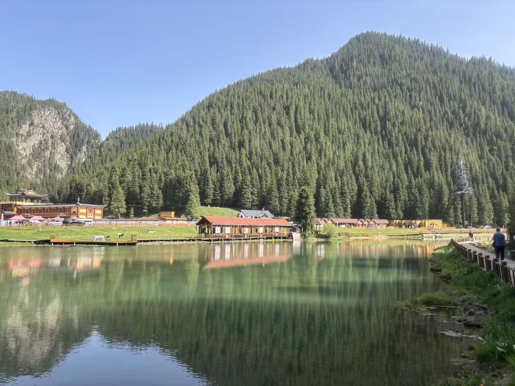 碧綠湖面平靜無波，完整倒映紅頂木屋與滿山雲杉。沿湖木棧道延伸向遠方，零星行人漫步觀景，巍峨山林作背景，建築融于山野，山水屋林相融，氛圍安靜柔和