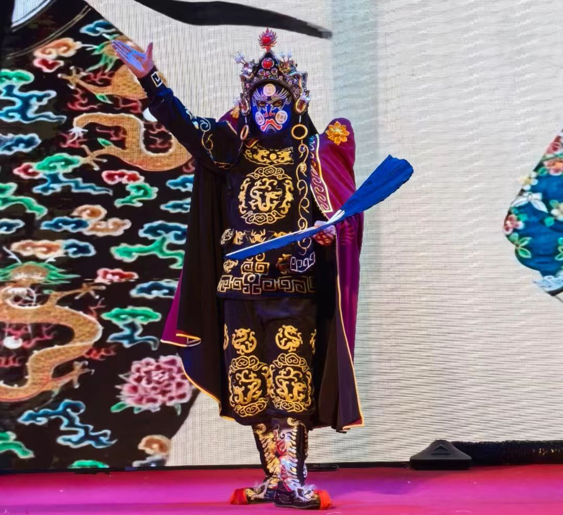 戲曲演員飾藍臉角色，穿黑金繡紋戲服，持藍扇展臂，背景繪龍紋，扮相華麗醒目