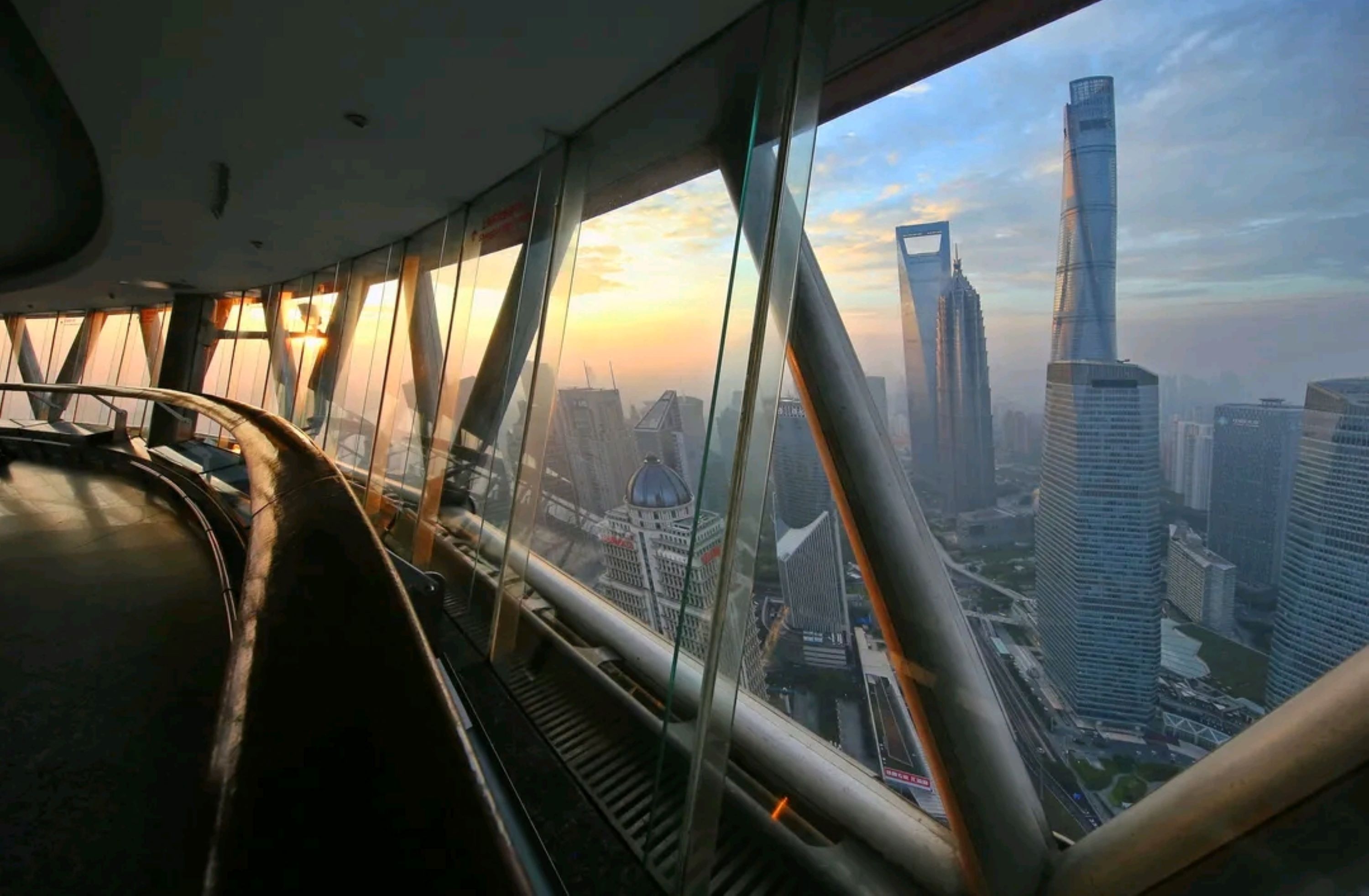 站在東方明珠高空環形觀景廊，全景落地窗將魔都風光盡收眼底。暖金色的晨光透過玻璃灑入廊道，窗外上海中心、環球金融中心等摩天大樓直插雲霄，雲海與城市天際線交融，盡顯高空俯瞰的壯闊與治癒