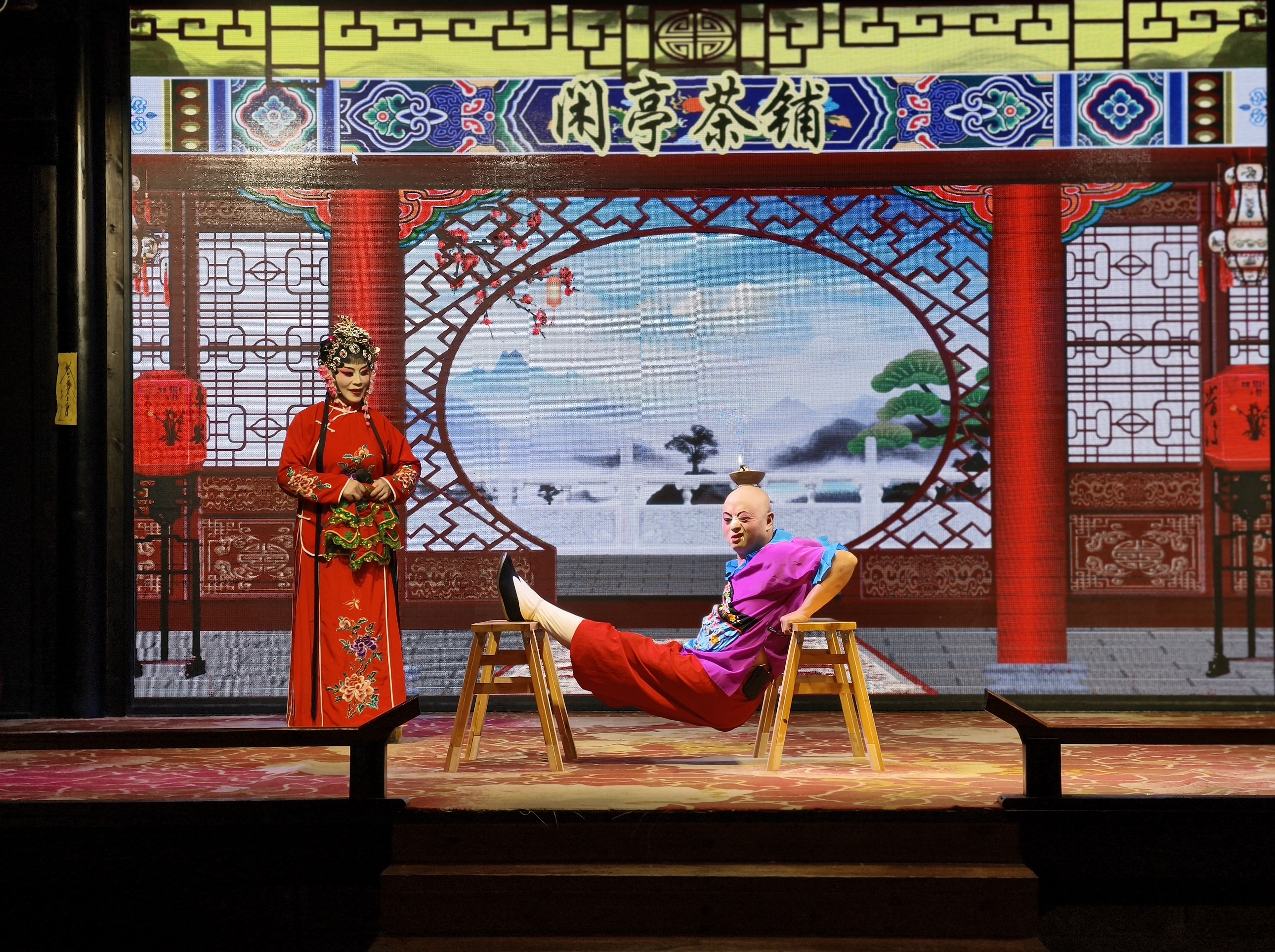 身著紅色戲服，頭戴華麗頭飾，立於一側；丑角演員則以高難度動作坐在兩把木凳之間，頭頂還燃著燈火，在 “閑亭茶鋪” 的佈景前，演繹出一段充滿詼諧與技藝的戲曲片段