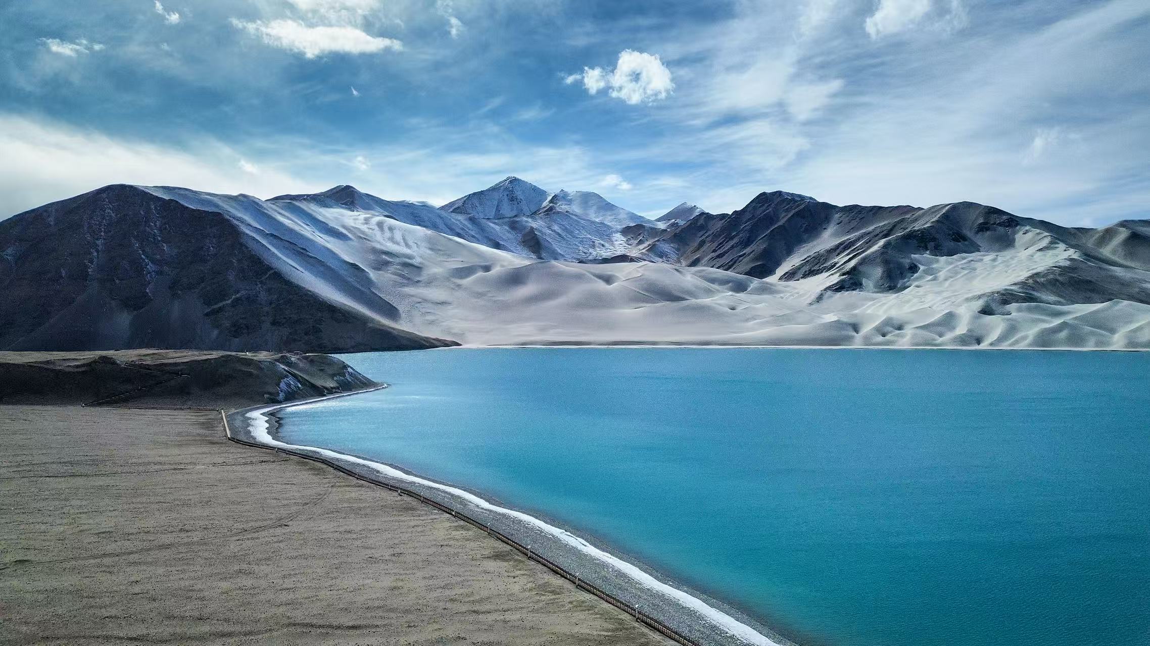 白沙湖的湖水呈現出夢幻的藍綠色，岸邊的白色沙丘如絲綢般柔滑，與遠處的雪山相映成趣。澄澈的藍天與白雲倒映在湖面，勾勒出一幅純淨的高原風光