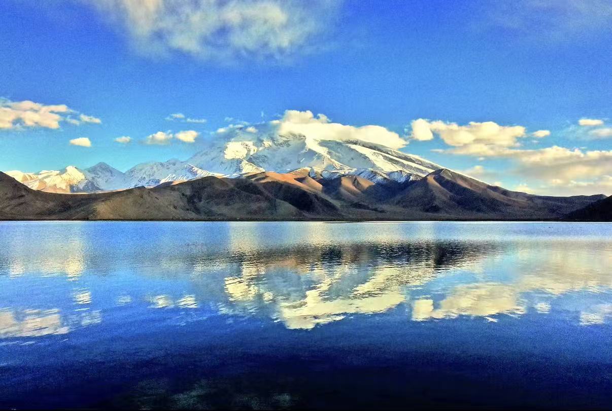 傍晚的慕士塔格峰被夕陽鍍上金邊，雪山與雲霞一同倒映在靜謐湖面，暖金色的柔光為冷峻的雪峰添了幾分溫柔，高原的寧靜與壯闊在此刻融為一體