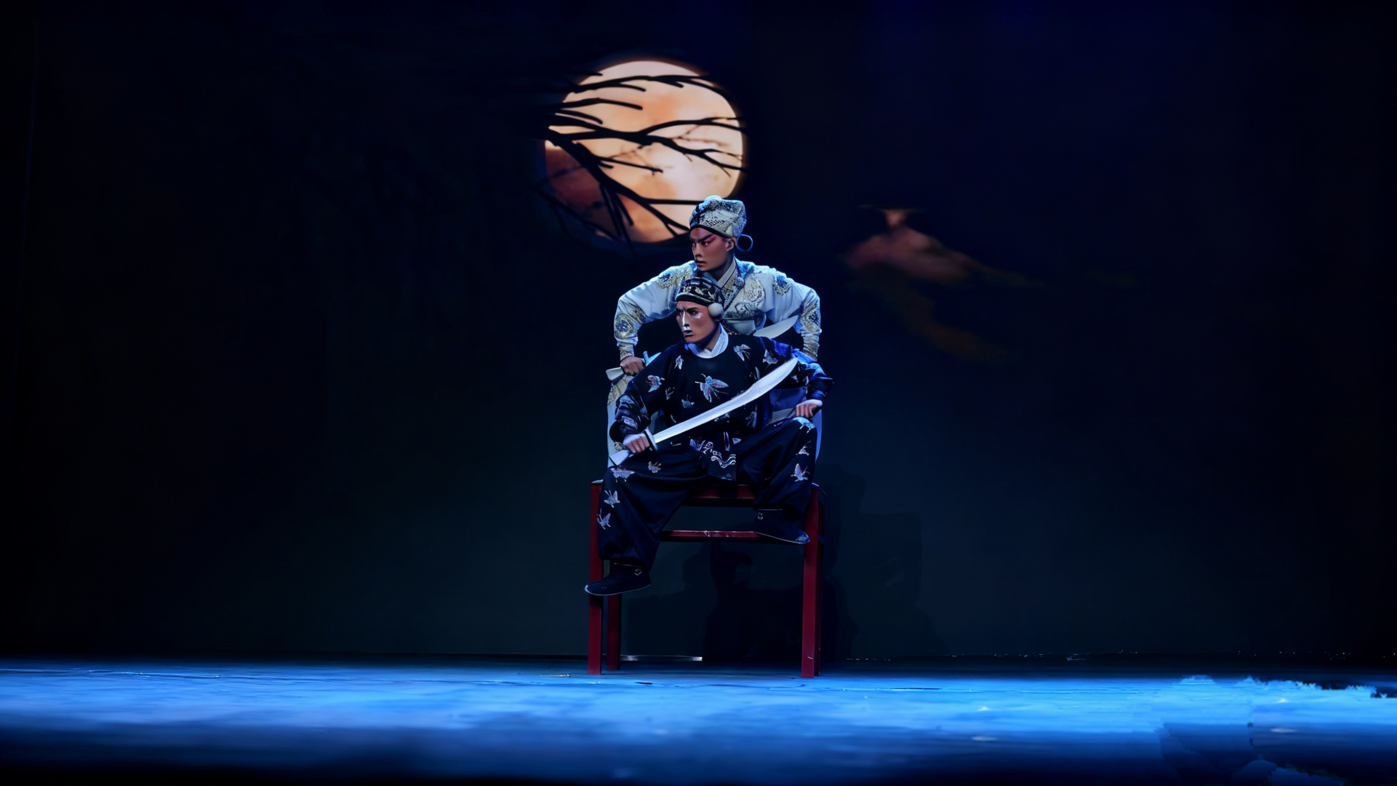 在月夜枯枝的佈景下,兩位京劇演員呈現出極具張力的畫面。一位演員,手持彎刀坐在紅色的椅子上;另一位演員身,立於其後,動作充滿壓迫感。將京劇武戲的緊張與懸疑感拉滿 在月夜枯枝的佈景下,兩位京劇演員呈現出極具張力的畫面。一位演員,手持彎刀坐在紅色的椅子上;另一位演員身,立於其後,動作充滿壓迫感。將京劇武戲的緊張與懸疑感拉滿