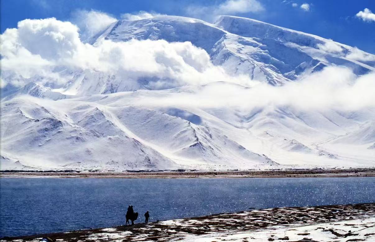 帕米爾高原喀拉庫勒湖畔的慕士塔格峰盛景。巍峨的雪峰被流雲輕擁，銀白山體在藍天映襯下聖潔巍峨。澄澈湖水泛著粼粼波光，岸邊一人一駝的剪影，為這片壯闊天地添上了一抹生動的人文氣息