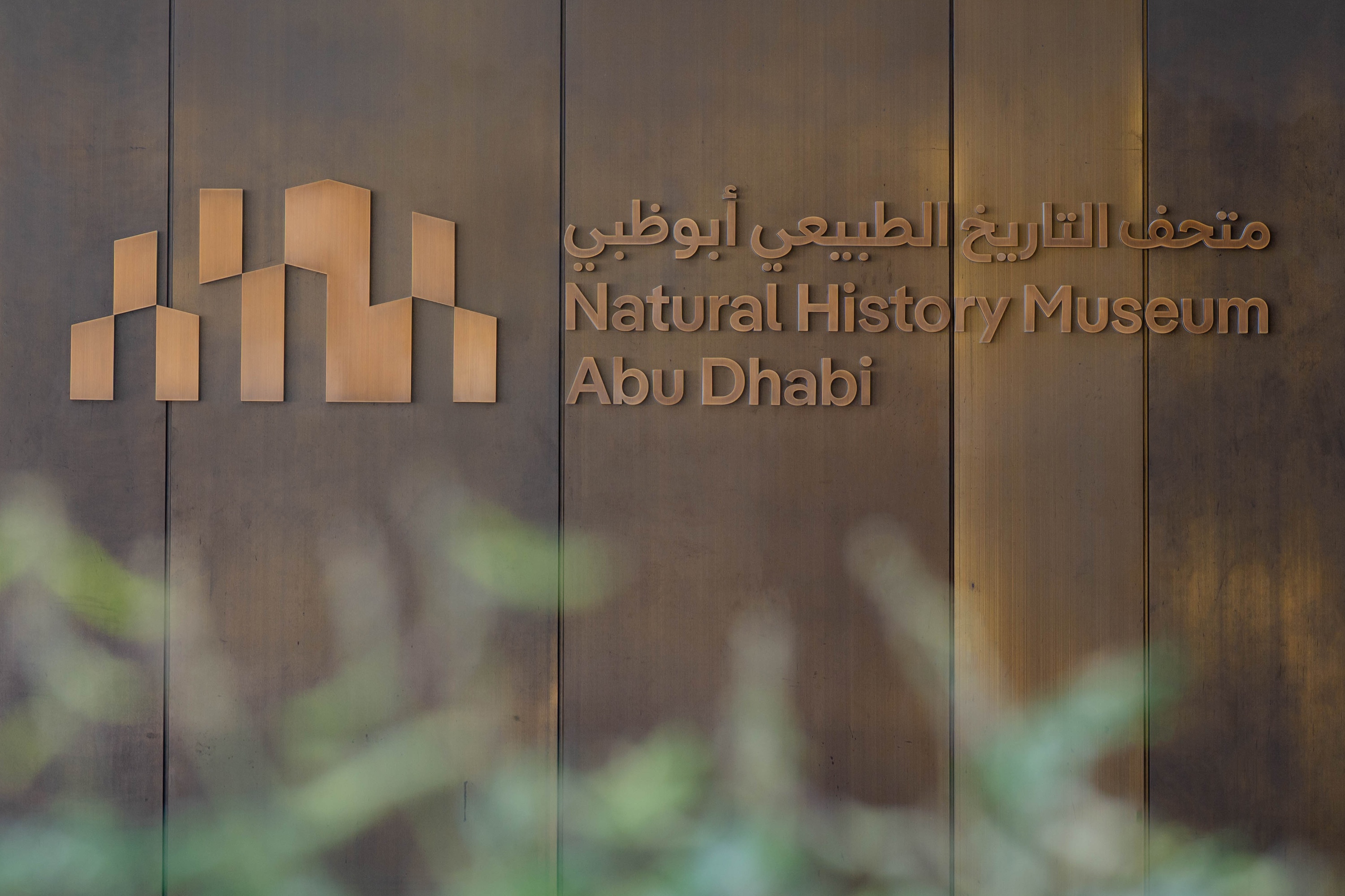 Bilingual signage and a bold bronze entrance welcome visitors to the Natural History Museum Abu Dhabi, celebrating knowledge, discovery, and Emirati heritage.
