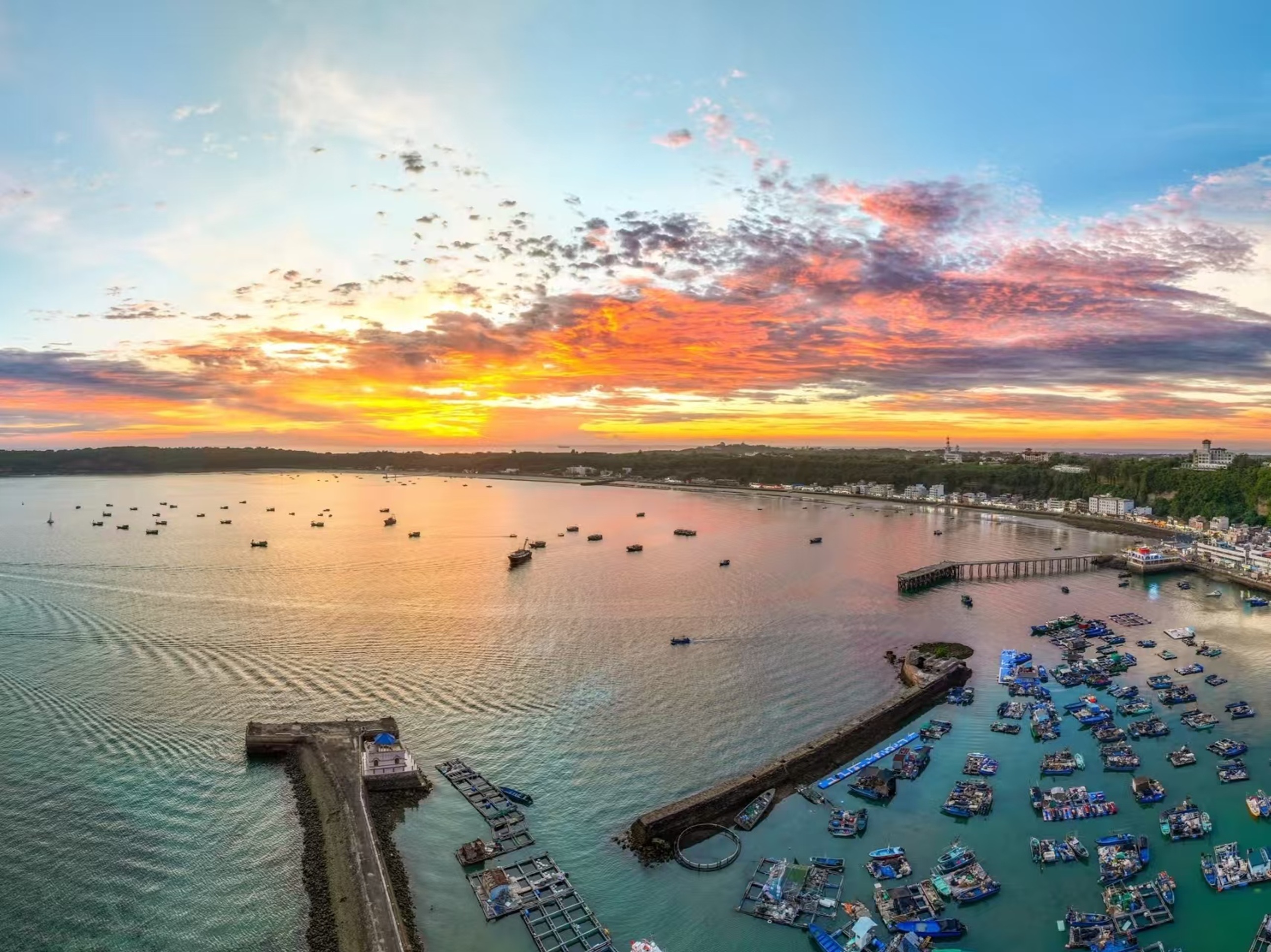 潿洲島碼頭的黃昏航拍,晚霞暈染出橙粉雲層,海面泛著暖光,數十艘漁船錯落停靠,岸邊村落與綠植相映,船痕劃破平靜水面,盡顯海島漁港的溫柔菸火氣 潿洲島碼頭的黃昏航拍,晚霞暈染出橙粉雲層,海面泛著暖光,數十艘漁船錯落停靠,岸邊村落與綠植相映,船痕劃破平靜水面,盡顯海島漁港的溫柔菸火氣