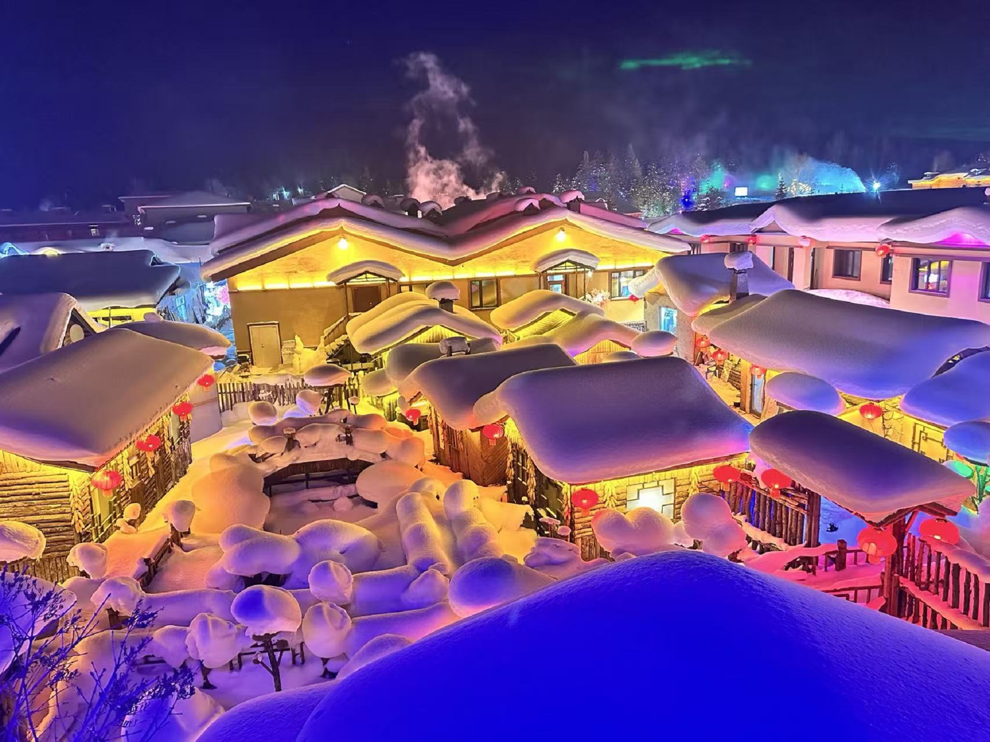 高空俯瞰雪鄉夜色，座座木屋頂著厚重雪蓋，紅燈籠錯落懸掛。暖黃燈光映亮木屋，造型圓潤的雪蘑菇遍佈房前屋後，遠處菸火升騰，盡顯東北雪鄉獨有的冬日浪漫
