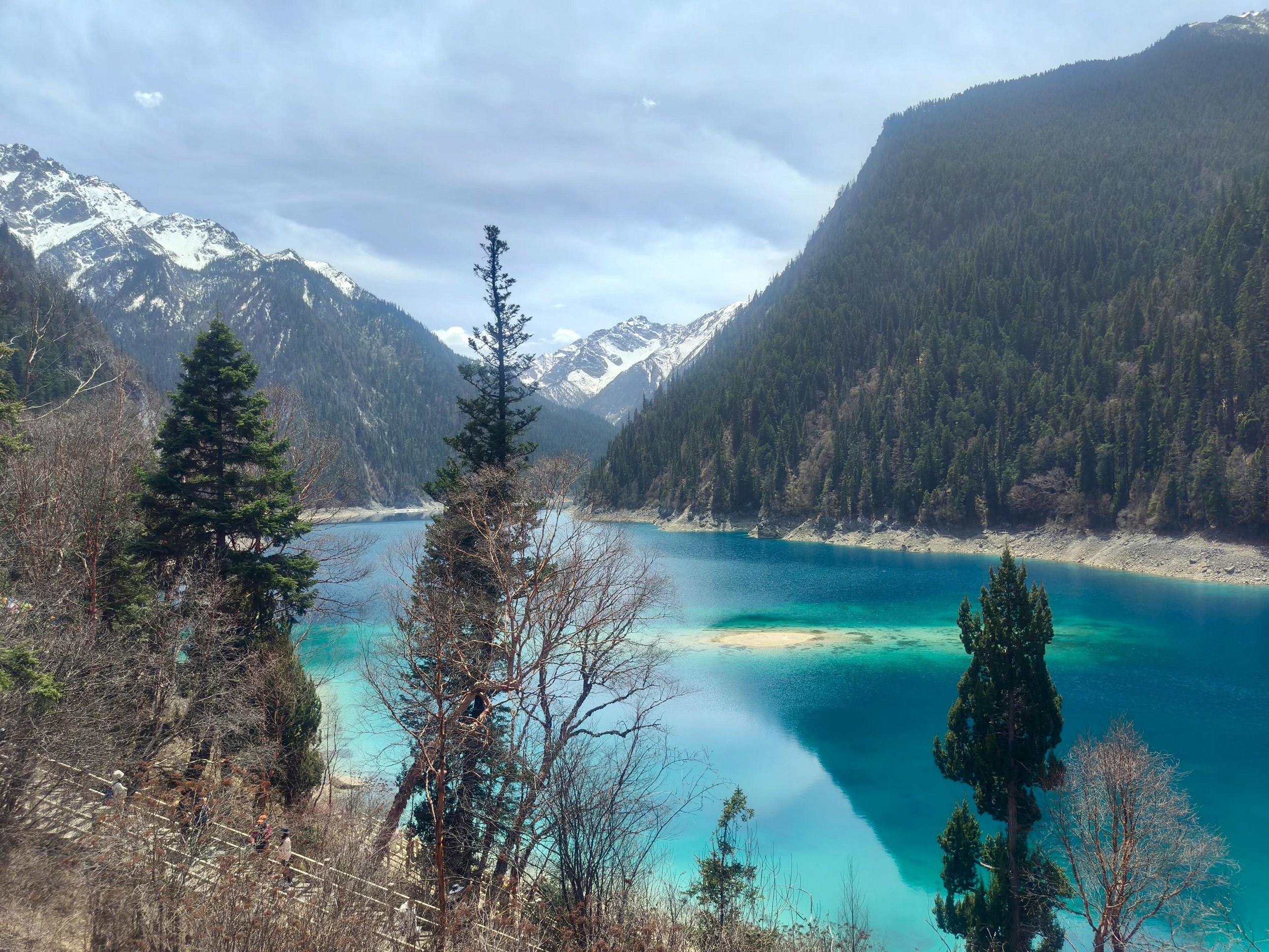 兩側山林巍峨聳立，蒼翠的冷杉與枯木錯落，中間的湖水像一條碧綠的玉帶蜿蜒在山谷間，藍綠交織的湖水清透見底，陽光灑在水面，波光粼粼間盡顯靈動