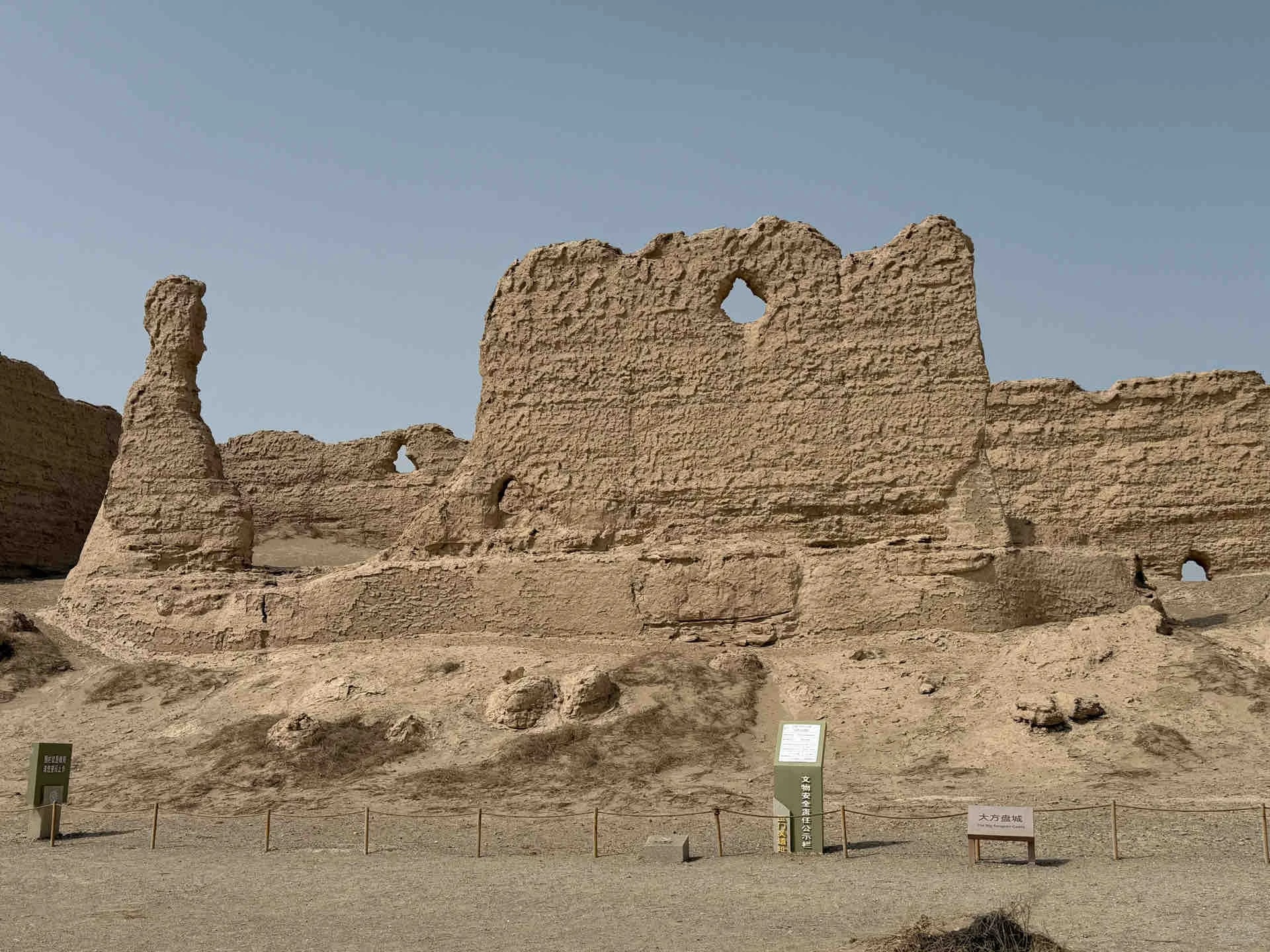 這是敦煌玉門關遺址中的大方盤城(河倉城)。畫面中,夯土築成的殘牆巍然屹立,牆體表面佈滿風蝕的溝壑與孔洞,顯露出歲月的滄桑痕跡。遺蹟整體呈不規則輪廓,部分坍塌的土塊與沙礫堆積腳下,映襯出大漠孤城的荒涼感。前景的資訊牌與圍欄提示這是受保護的歷史遺存。 這是敦煌玉門關遺址中的大方盤城(河倉城)。畫面中,夯土築成的殘牆巍然屹立,牆體表面佈滿風蝕的溝壑與孔洞,顯露出歲月的滄桑痕跡。遺蹟整體呈不規則輪廓,部分坍塌的土塊與沙礫堆積腳下,映襯出大漠孤城的荒涼感。前景的資訊牌與圍欄提示這是受保護的歷史遺存。