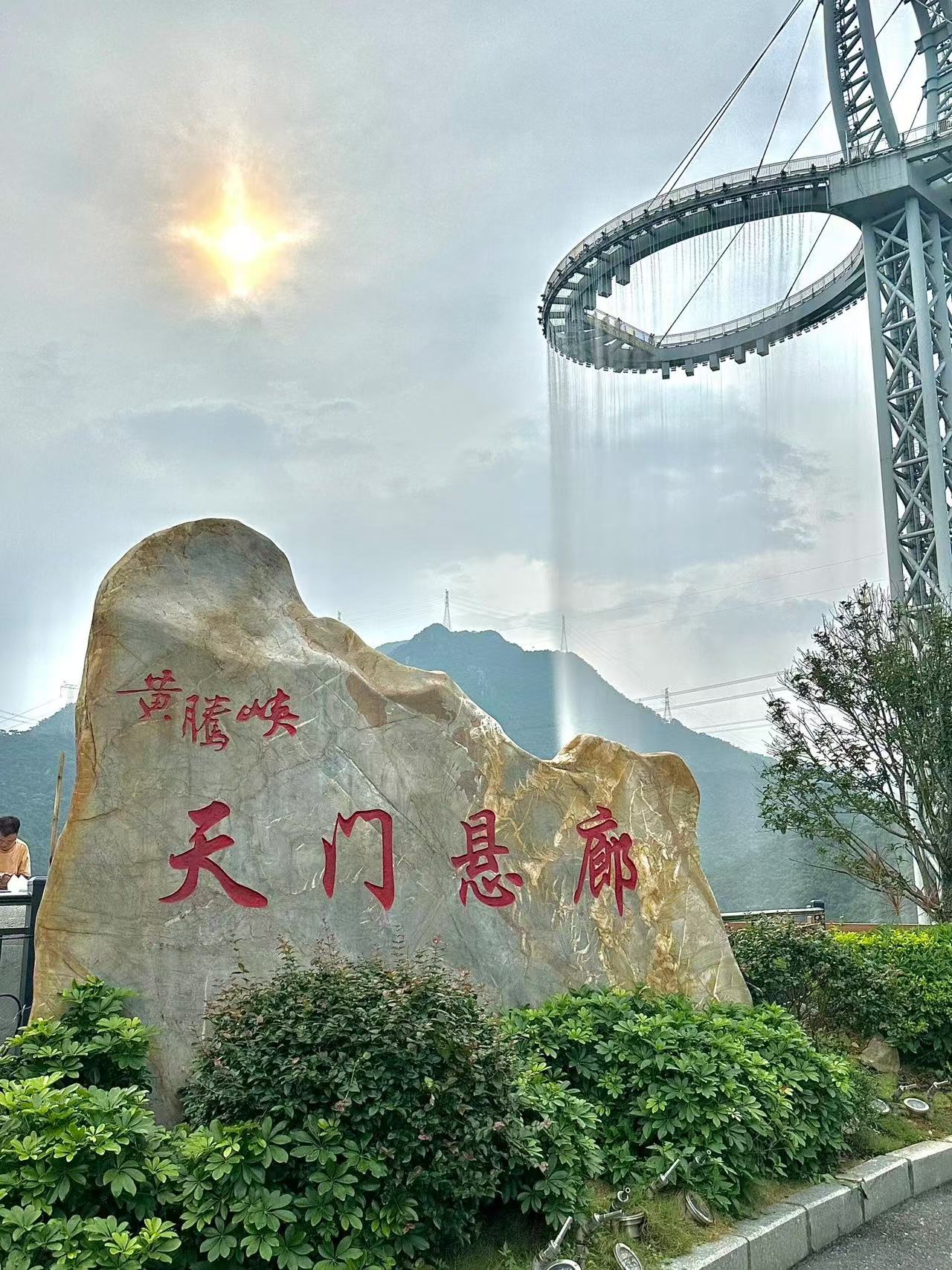 天門懸廊打卡地標！巨型石碑刻著 “黃騰峽 天門懸廊”，背景是震撼的環形玻璃懸廊，瀑布從廊頂傾瀉而下，陽光穿透雲層，氛圍感與打卡感雙在線