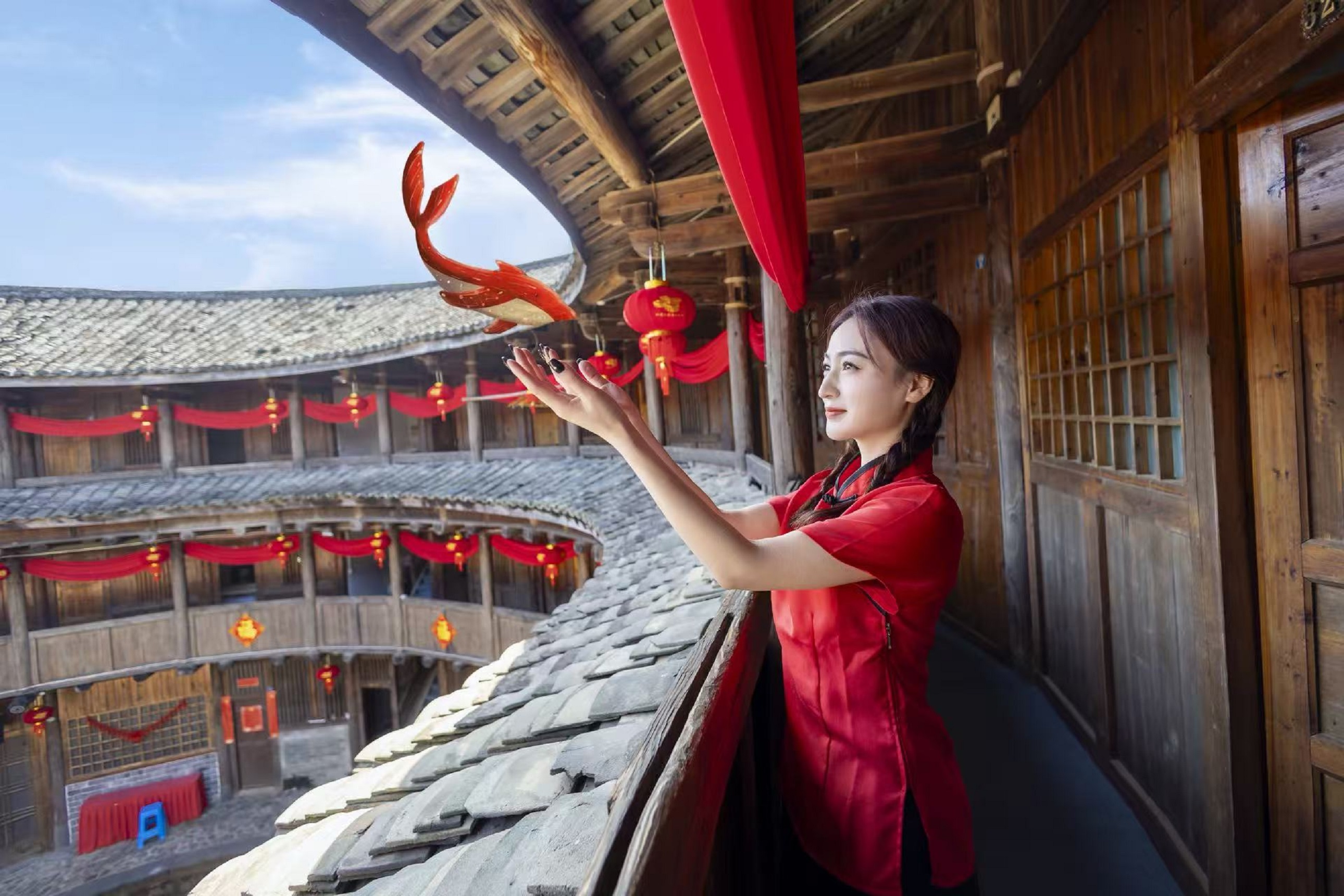 紅裝的姑娘探身伸手,指尖輕觸懸在簷下的紅魚紙鳶,土樓層層疊疊的木質結構在身後鋪展 紅裝的姑娘探身伸手,指尖輕觸懸在簷下的紅魚紙鳶,土樓層層疊疊的木質結構在身後鋪展