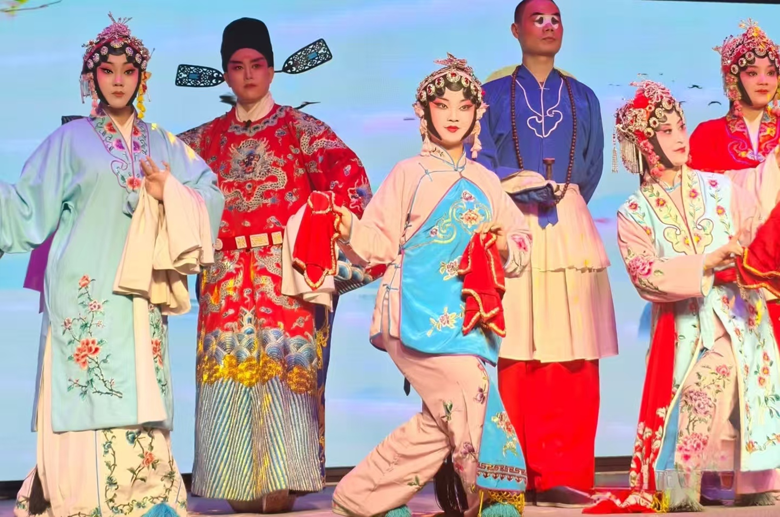 多位戲曲演員同臺：旦角衣飾繡花鳥，生角著紅龍袍，丑角扮相詼諧，整體陣容明豔鮮活