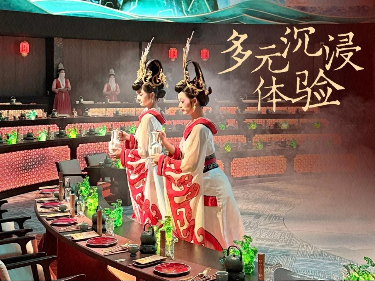 兩位侍女身著白底紅紋的唐風寬袖襦裙，頭戴華麗金飾高髻，手持酒壺立於長桌旁，姿態優雅如古畫中走出的仕女；席間長桌鋪陳著紅釉餐具、青綠器皿，搭配古式茶具，將用餐器具與國風美學融為一體。