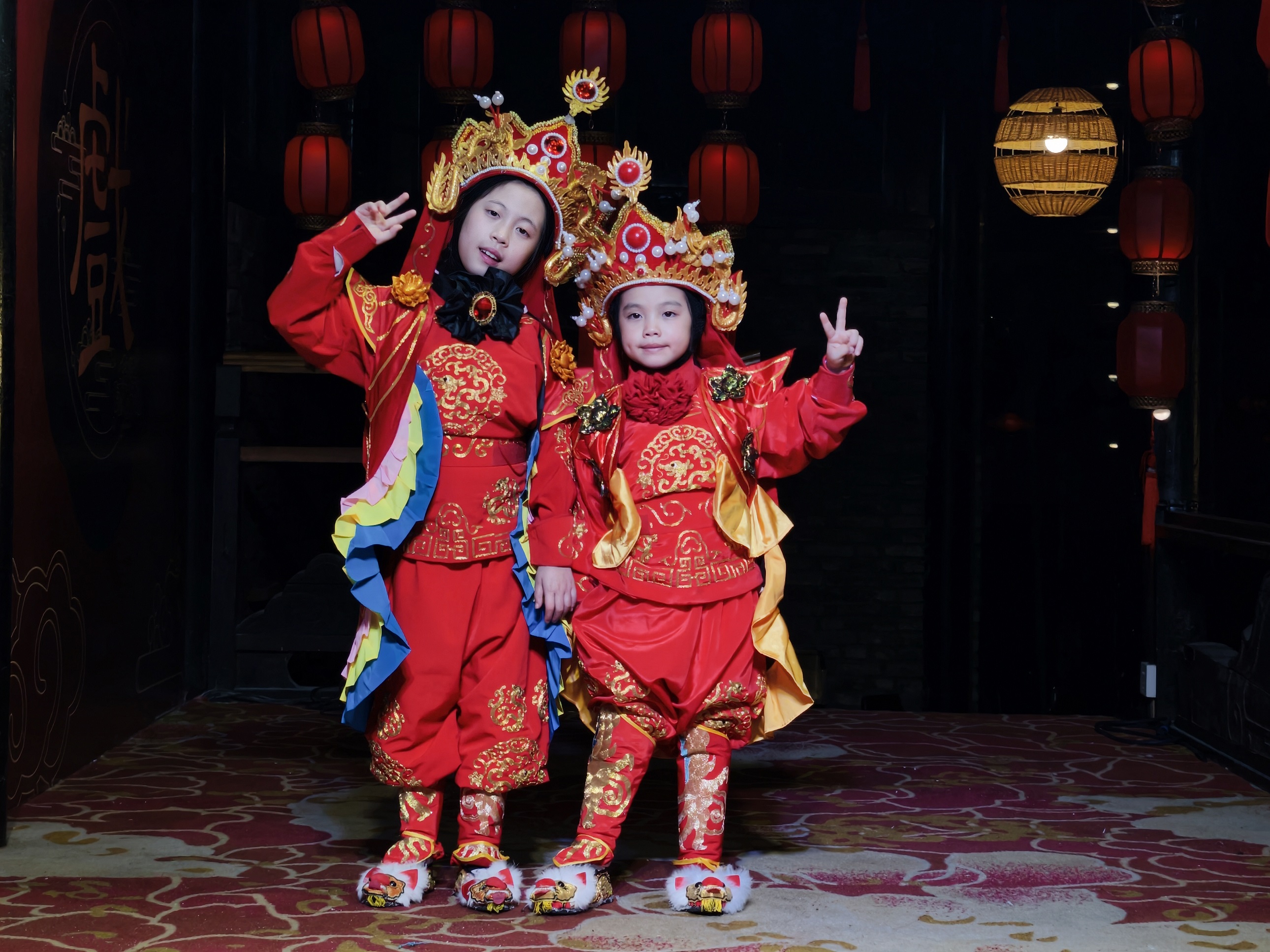 充滿傳統氛圍的場景中，兩個孩童身著華麗的紅色戲服，頭戴精緻的戲曲盔頭，在紅燈籠的背景下，俏皮地比出 “耶” 的手勢，童真與傳統戲曲元素碰撞出別樣的可愛畫面