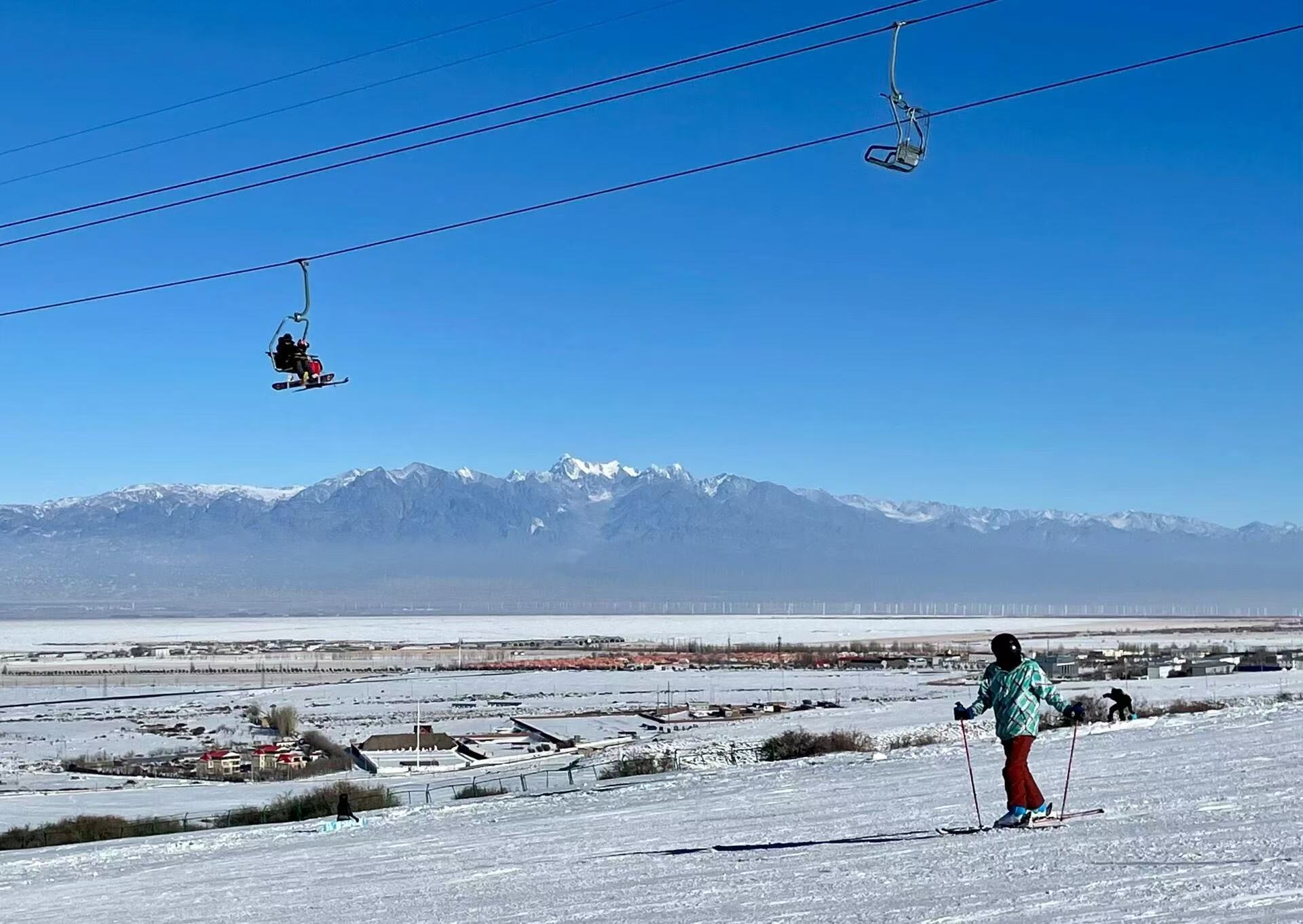 晴朗藍天之下，滑雪者持雙杖滑行雪坡，上空纜車搭載遊客穿行索道。遠方巍峨雪山連綿橫亙，山前是覆雪平原與村落建築，遠景層次豐富，雪山、雪場、城鎮同框，視野開闊壯美