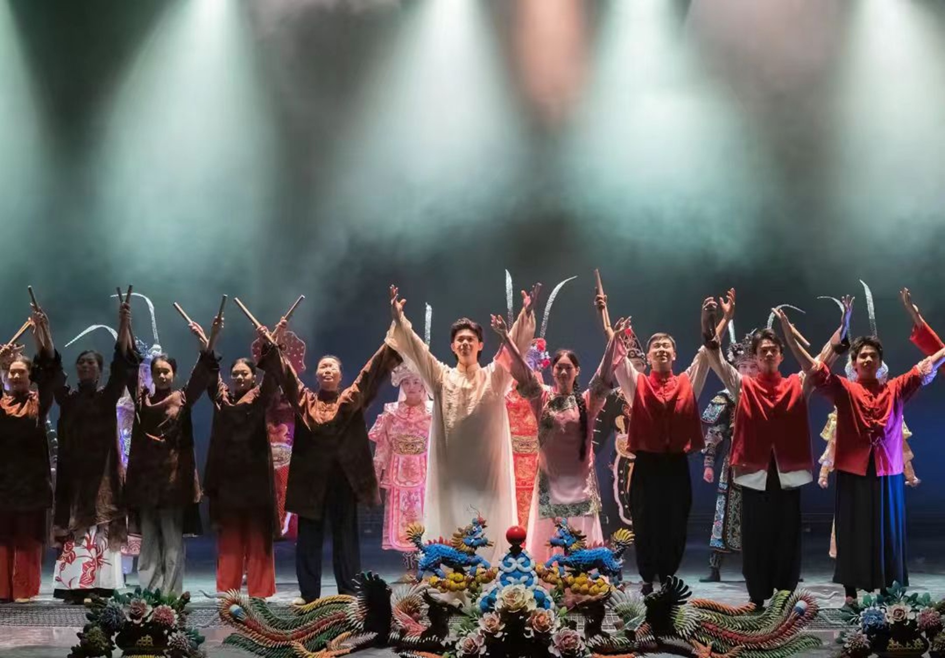 演員們著傳統服飾並肩展臂，臺前龍鳳裝飾亮眼，燈光朦朧顯舞臺謝幕溫情