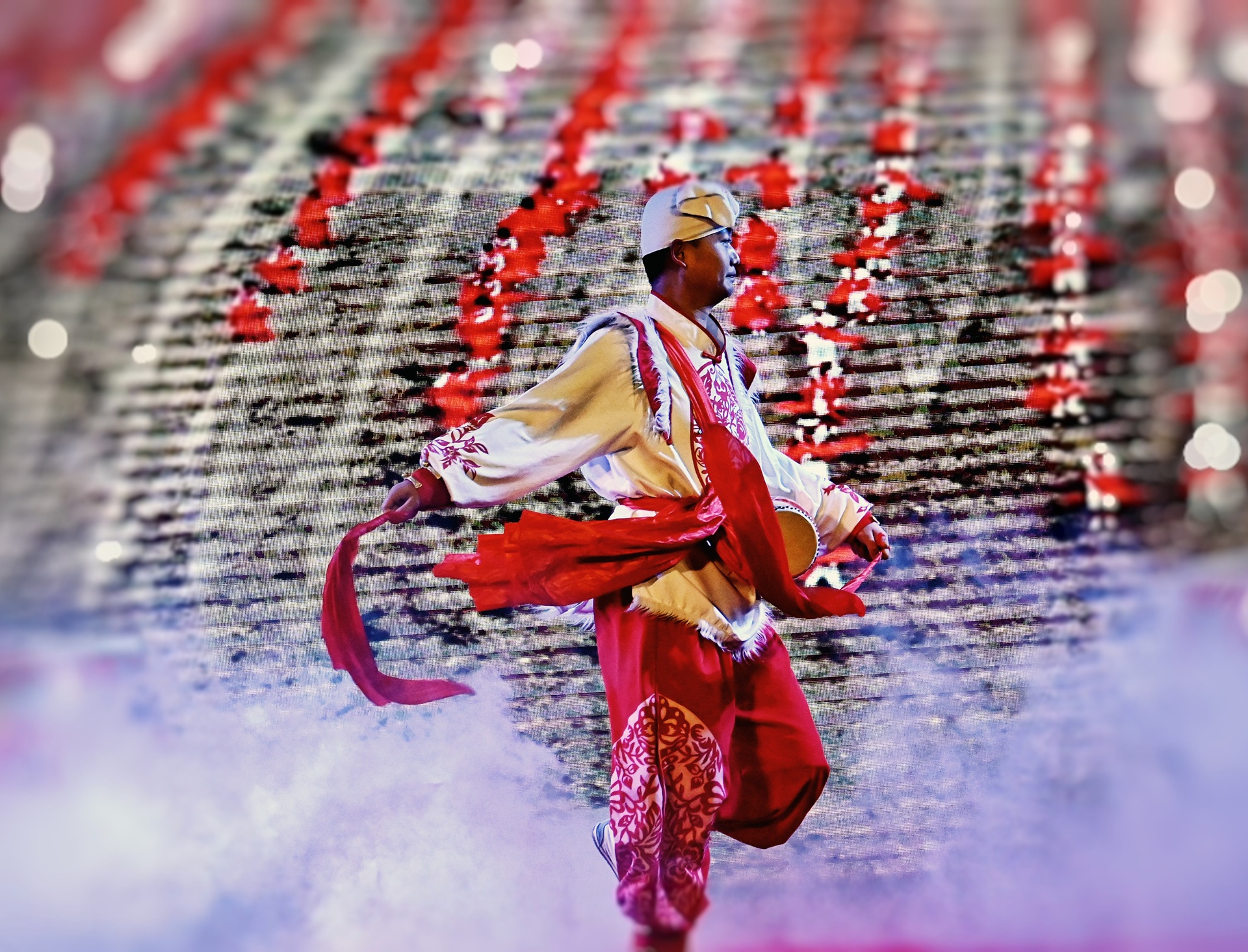 舞者穿白紅民族服飾，揮紅綢帶，背景是梯隊表演者，菸霧繚繞，盡顯民俗舞蹈的熱烈