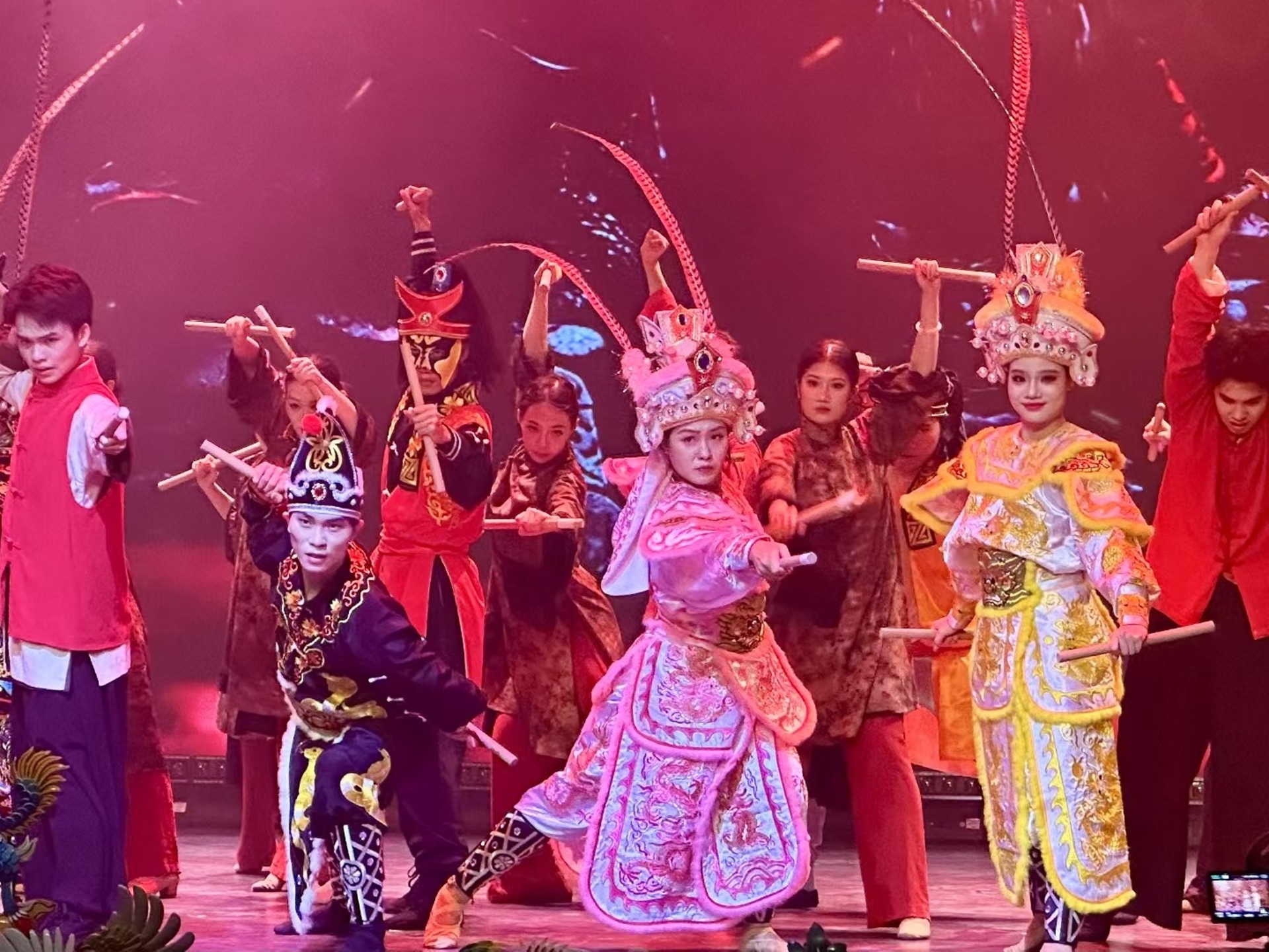 演員們持棍伴戲曲裝扮，粉黃戲服配紅調燈光，動作利落顯表演張力