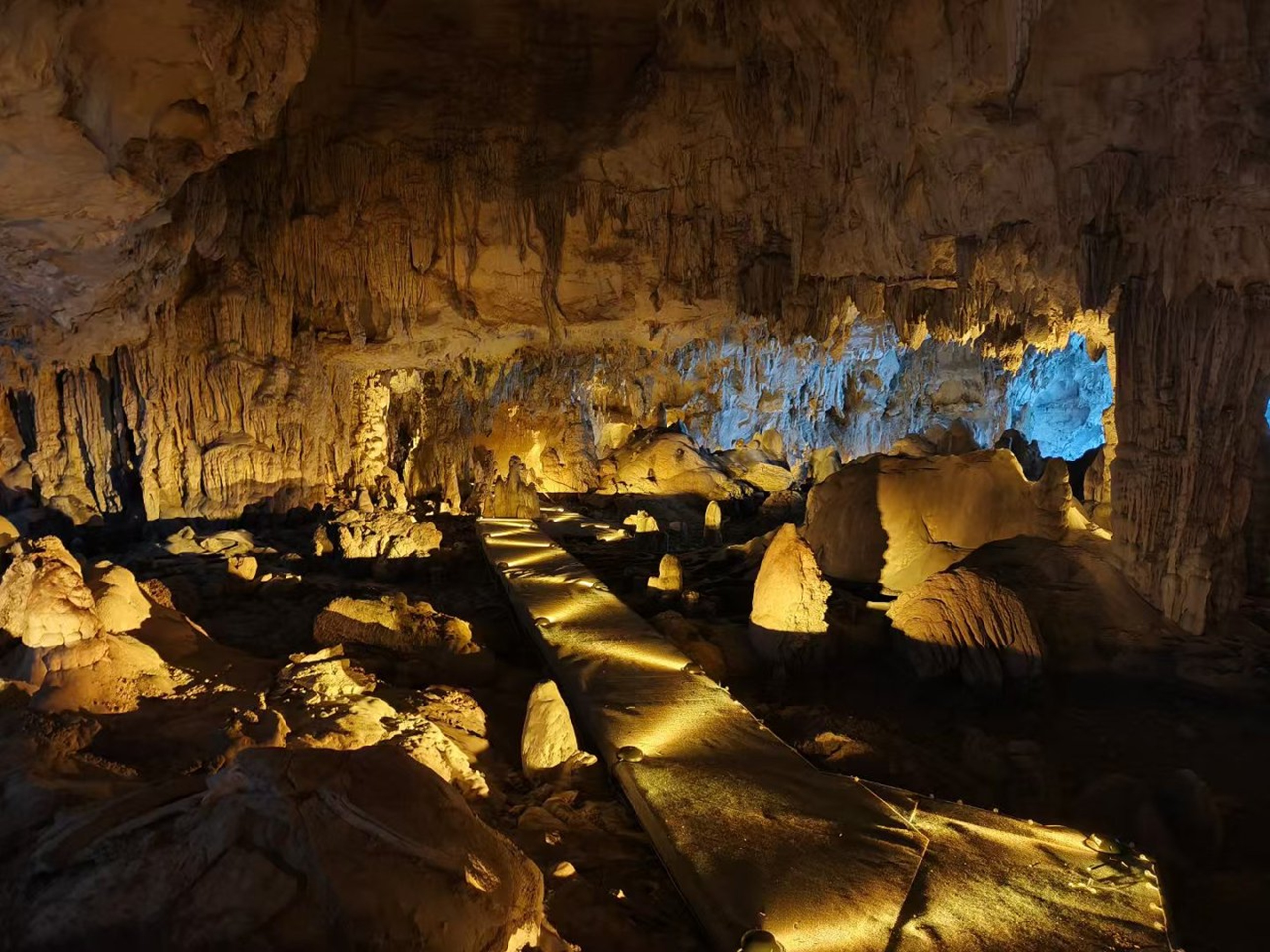 溶洞內步道延伸向深處,暖黃與冷藍燈光交錯,鐘乳石如垂簾般掛滿巖壁,彷彿踏入時光凝固的地心秘境 溶洞內步道延伸向深處,暖黃與冷藍燈光交錯,鐘乳石如垂簾般掛滿巖壁,彷彿踏入時光凝固的地心秘境