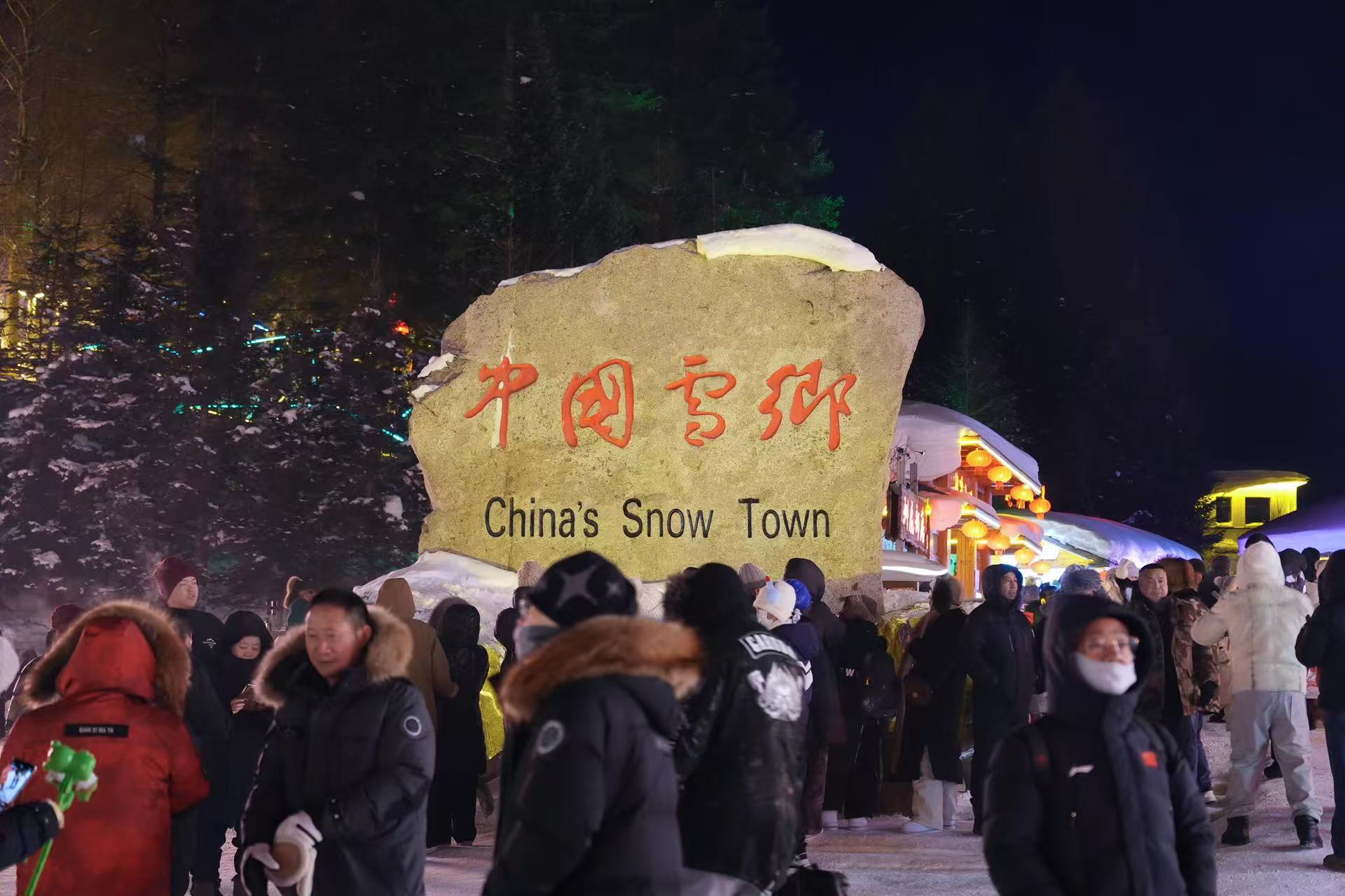 夜幕下中國雪鄉石碑醒目矗立，紅字刻石搭配英文標識，四周遊人絡繹不絕。周邊紅燈籠燈火璀璨，夜色與皚皚白雪相映，遊客駐足打卡，記錄來到雪鄉的標誌性時刻，熱鬧的冬日旅行氛圍感拉滿
