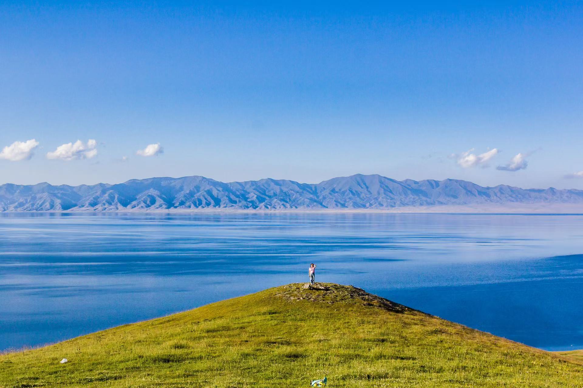 青綠緩坡草丘伸向湖畔，一人獨立坡頂遠眺賽里木湖。湖面鋪展層層漸變藍，遠方天山山脈輪廓柔和，幾縷白雲浮於晴空，遼闊湖山盡收眼底，開闊遼闊，氛圍感自由鬆弛