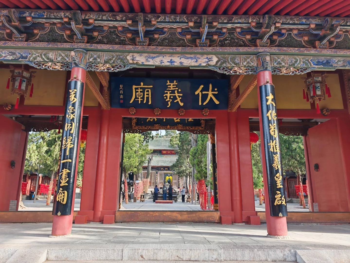 這是甘肅天水伏羲廟的正門——欞星門。畫面中，古典廟宇門樓莊嚴矗立，重簷歇山頂覆以傳統瓦飾，斗拱層疊彰顯明清建築精髓。門楣懸“伏羲廟”黑底金字匾額，筆力遒勁，兩側黑柱鐫刻“律令黔黎一畫開”，頌揚伏羲創制八卦、肇啟文明的歷史功績。硃紅立柱與斑斕彩繪相映，簷下木雕隱約可見神話紋樣，傳遞著上古文化的神秘氣息。作為中華人文始祖伏羲的祭祀聖地，這座廟宇以凝重的建築語言詮釋著華夏文明的起源，成為傳承根脈文化、凝聚民族認同的重要象徵。