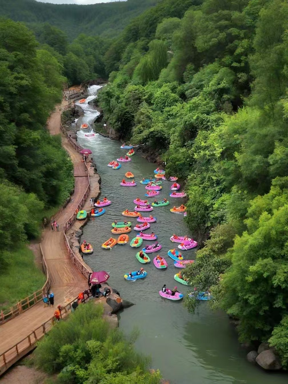全景視角看黃騰峽漂流！峽谷河道里，五顏六色的皮筏艇順流而下，兩岸青山如黛、綠樹成蔭，蜿蜒木道旁遊客往來，盡顯漂流的熱鬧與山野的治癒