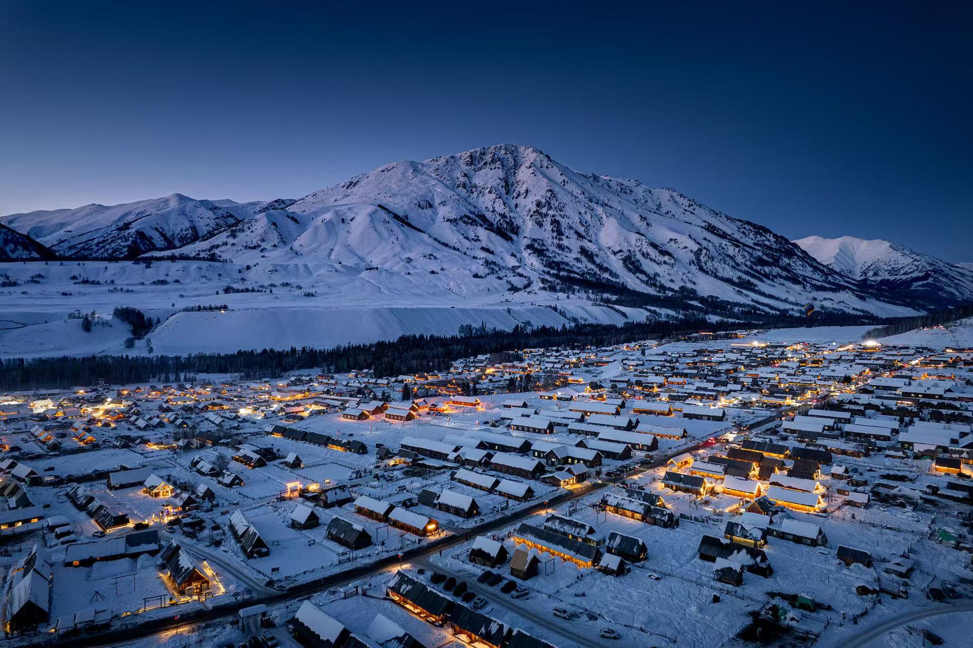 夜幕下禾木村落燈火點點，木屋覆滿厚雪。巍峨雪山矗立身後，冷暖色調交融，靜謐夢幻，像冬日童話小鎮