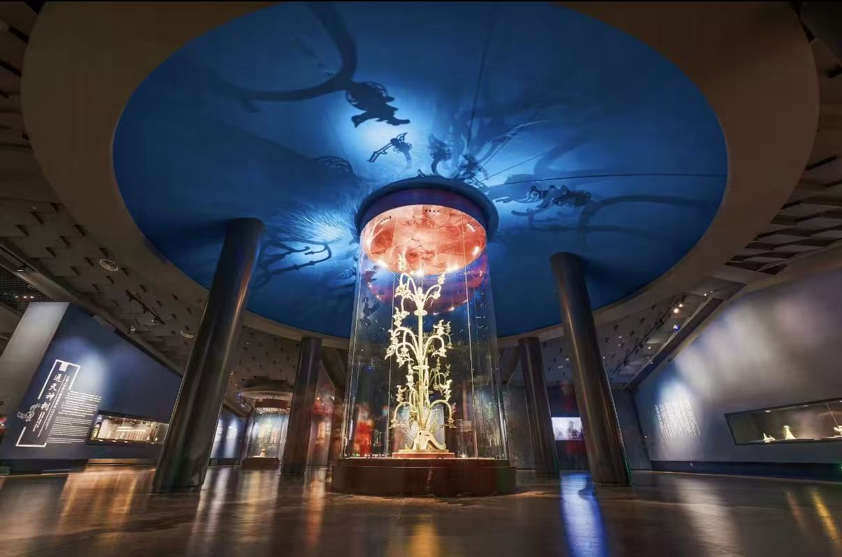 三星堆博物館的青銅神樹被置於玻璃展櫃中,金色枝幹舒展,造型神秘奇幻。穹頂藍色光影投射神鳥剪影,營造出古蜀通天神話的莊重氛圍 三星堆博物館的青銅神樹被置於玻璃展櫃中,金色枝幹舒展,造型神秘奇幻。穹頂藍色光影投射神鳥剪影,營造出古蜀通天神話的莊重氛圍