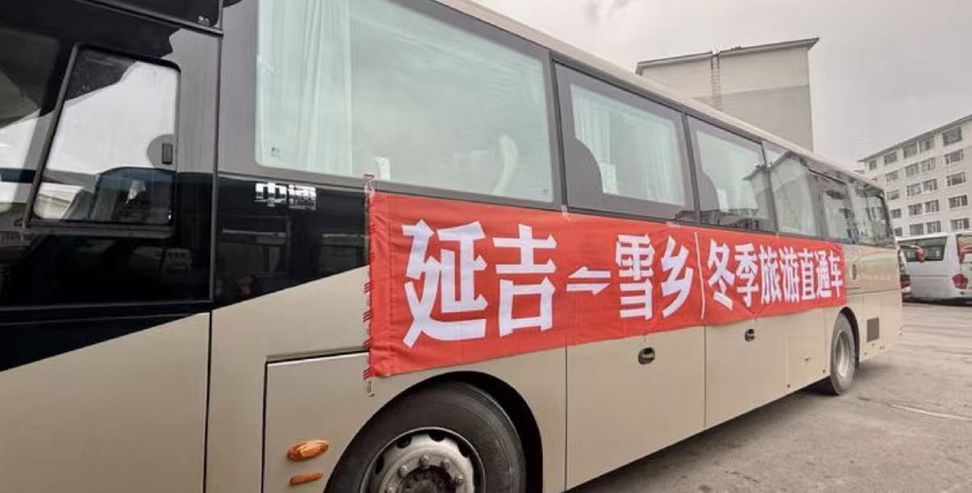 車身為淺米色旅遊大巴，車身側面懸掛醒目紅色橫幅，印有 “延吉⇋雪鄉冬季旅遊直通車” 白色大字。底層配備大容量行李艙，是雪鄉往返延吉、長白山跨線營運車輛，在雪鄉景區牡丹江東集站點上下客